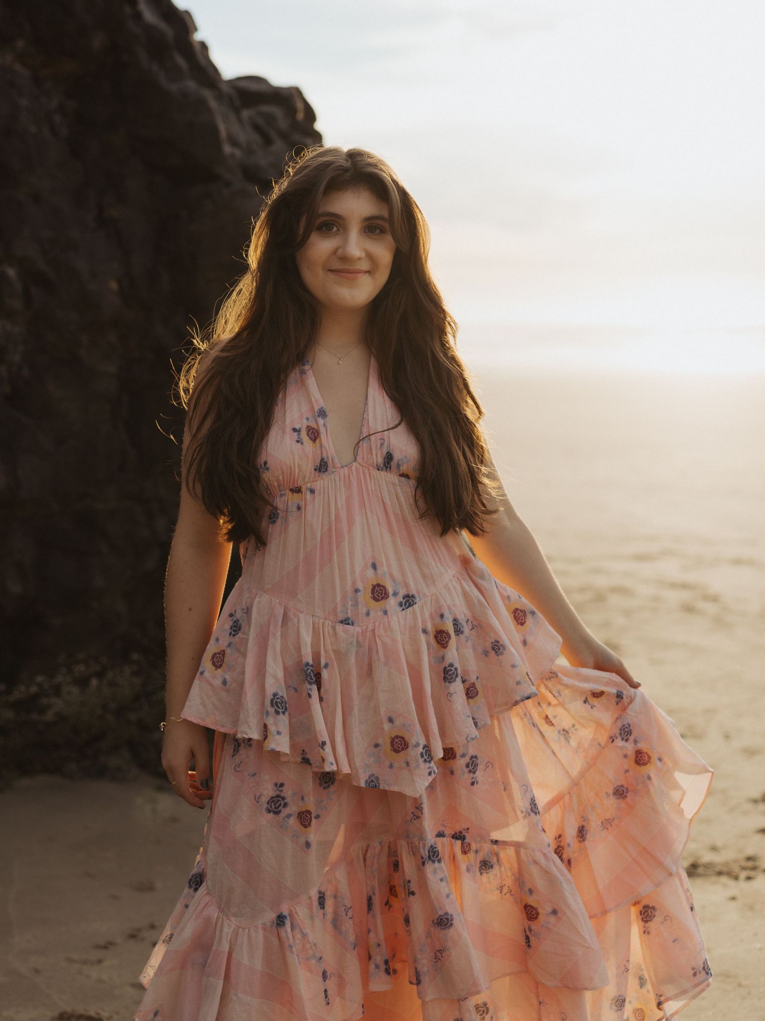 Young woman in pink floral tiered dress posing on a rocky beach at sunset with flowing dark hair. Oregon Coast senior Photographer.