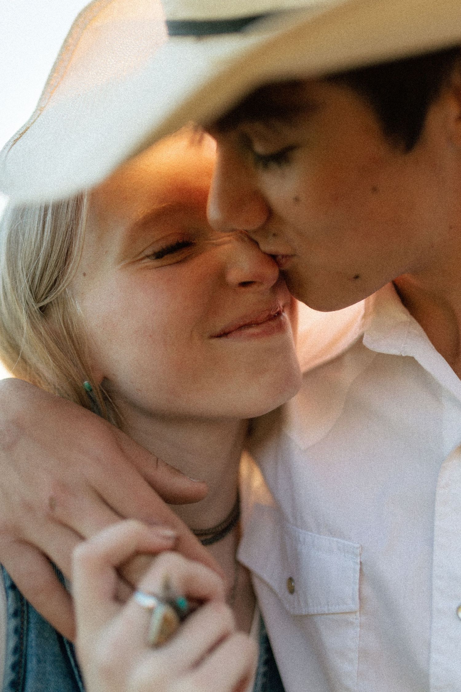 Romantic couple embracing at sunset, man in cowboy hat kissing woman on cheek in soft golden light.