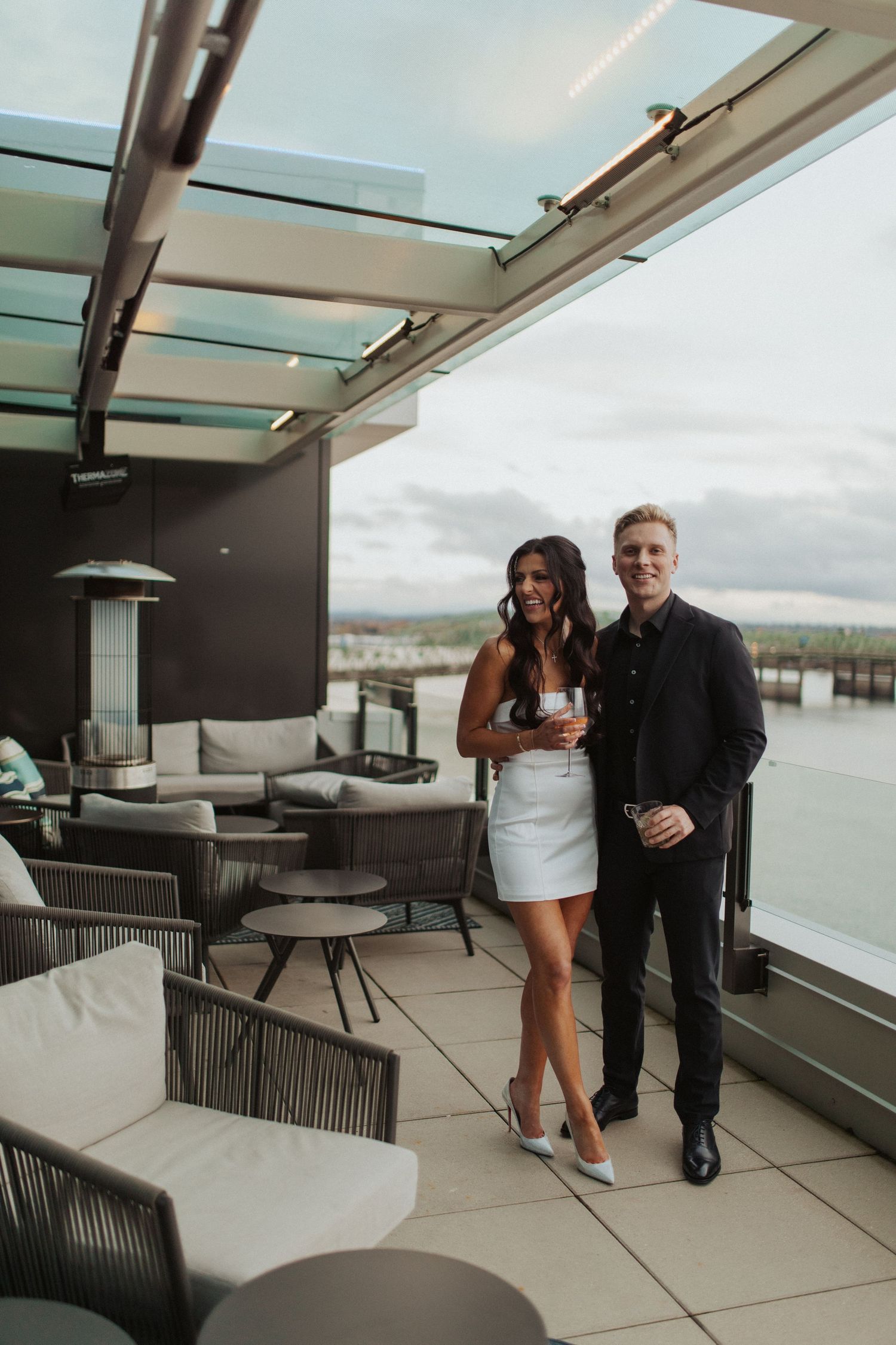 Stylish couple smiling on a rooftop terrace with waterfront views, woman in white mini dress holding champagne.