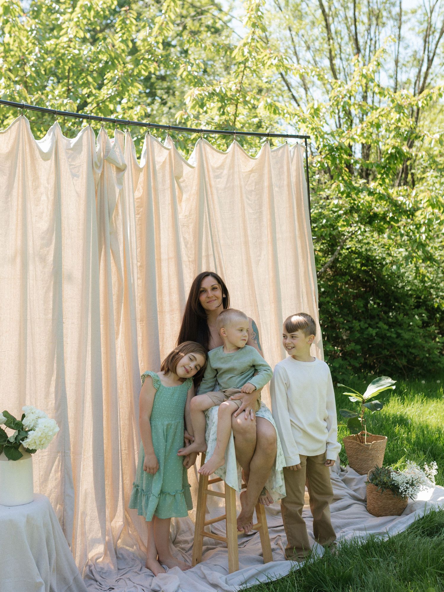 A mother poses with her three children in front of a draped curtain backdrop in a sunny garden.