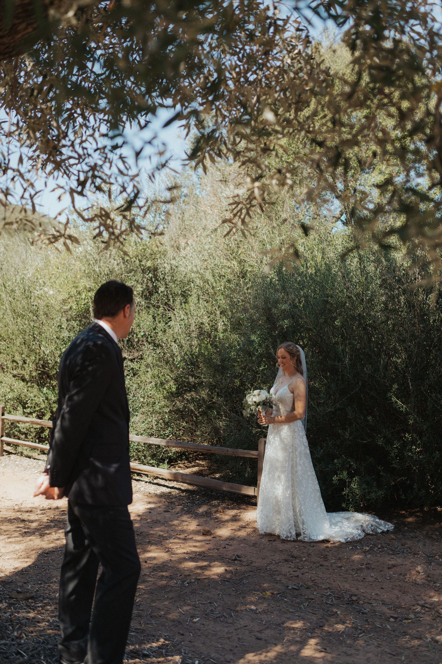 Bride in lace gown holds white bouquet during first look with groom in black suit outdoors.