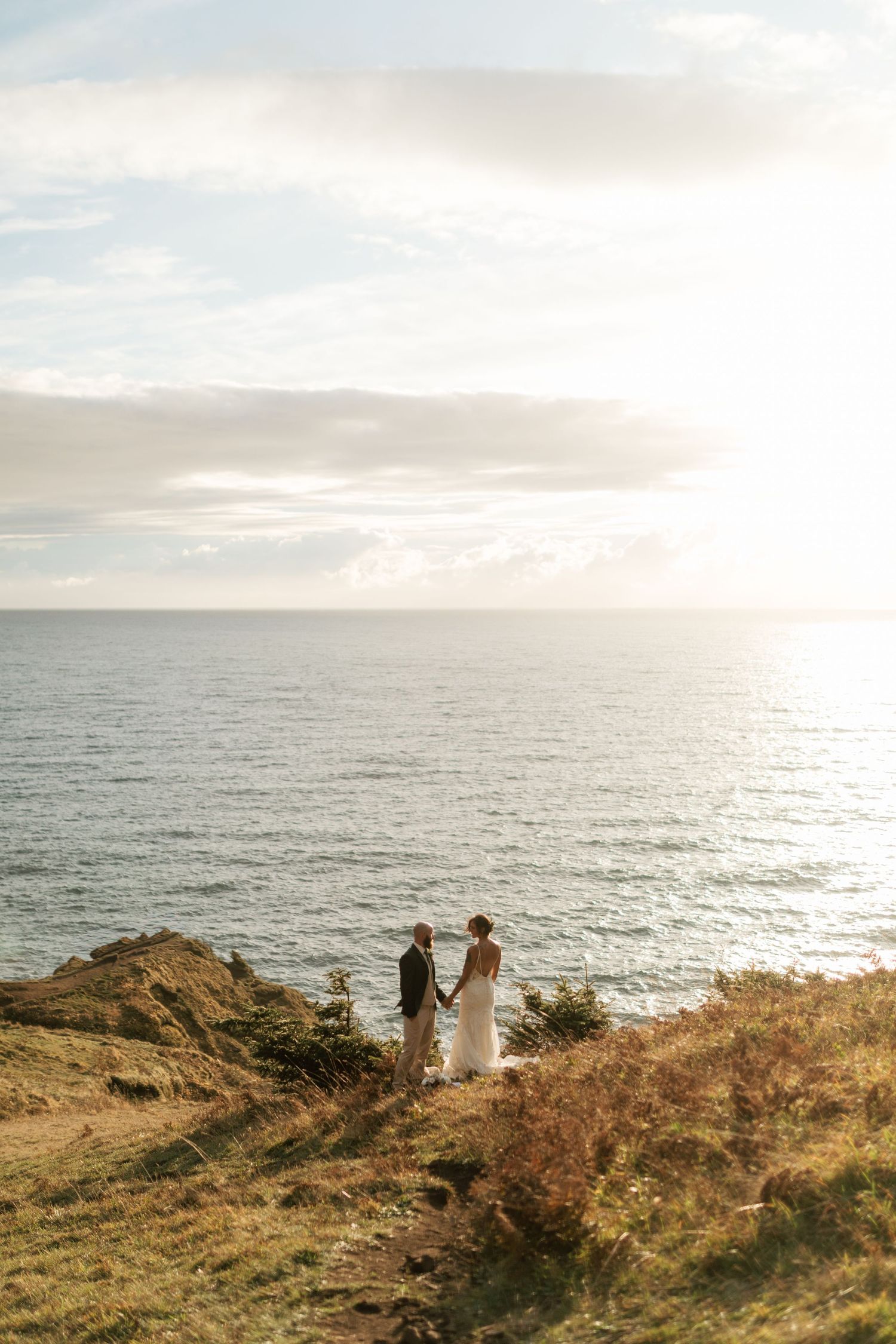 Bride and groom hold hands on a coastal cliff overlooking a sunlit ocean horizon.
