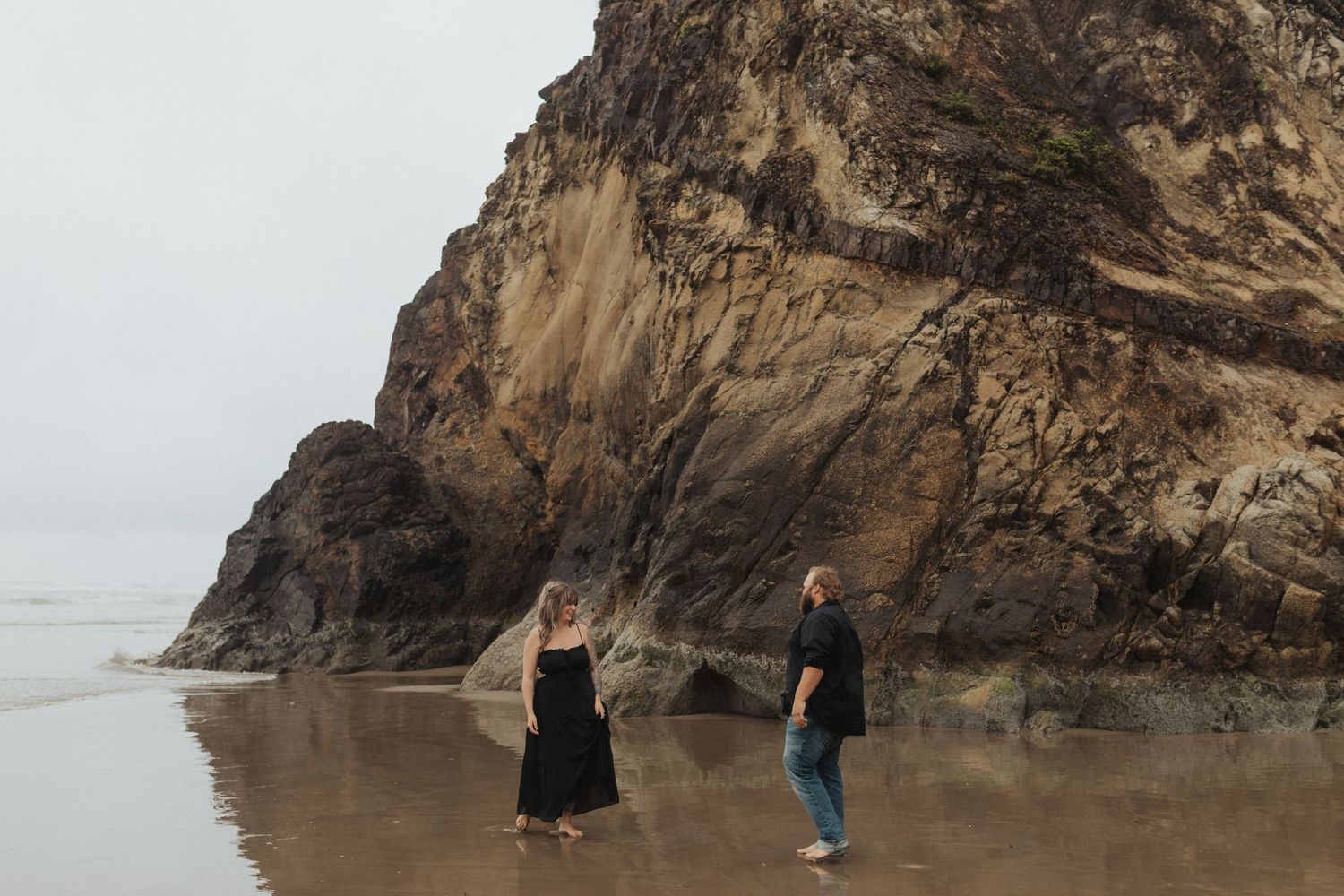 Couple in matching black outfits walking on reflective wet sand near dramatic rocky cliffs.