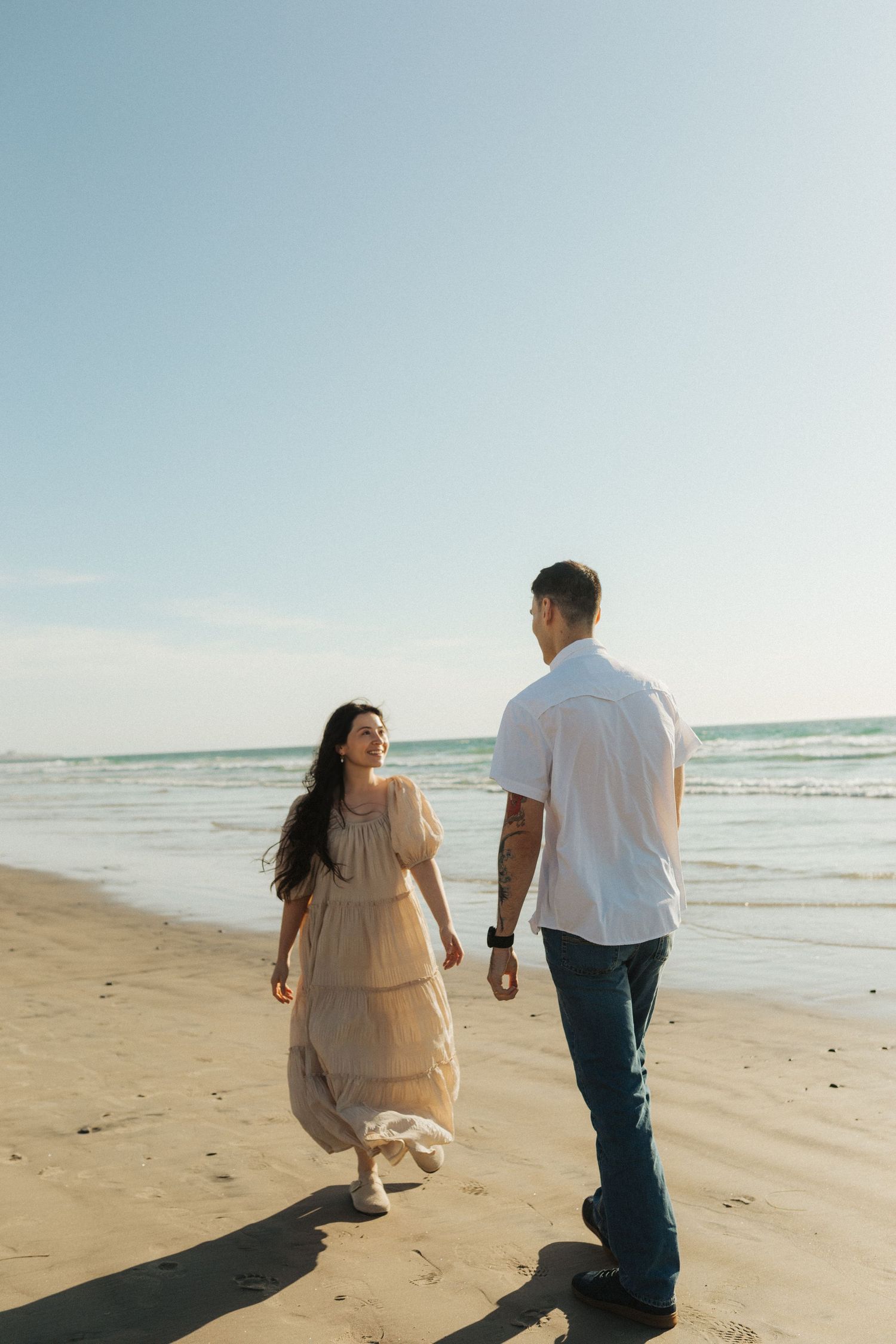 Couple walking hand in hand along a sunny beach, woman in flowing beige dress with ocean waves behind them.