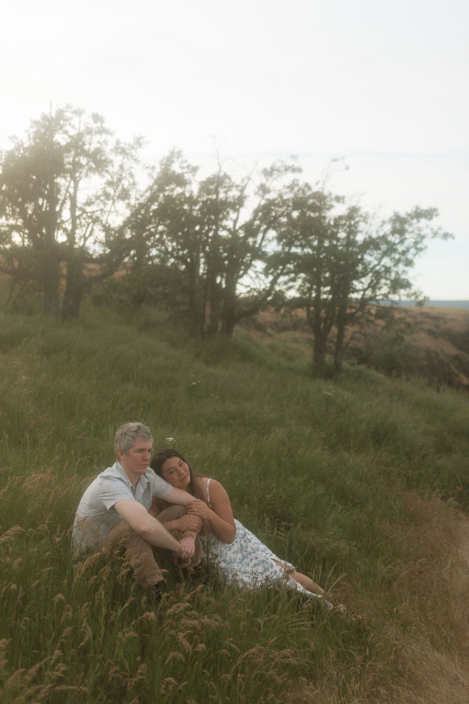 Couple sitting closely together on a grassy hillside at golden hour, leaning on each other.