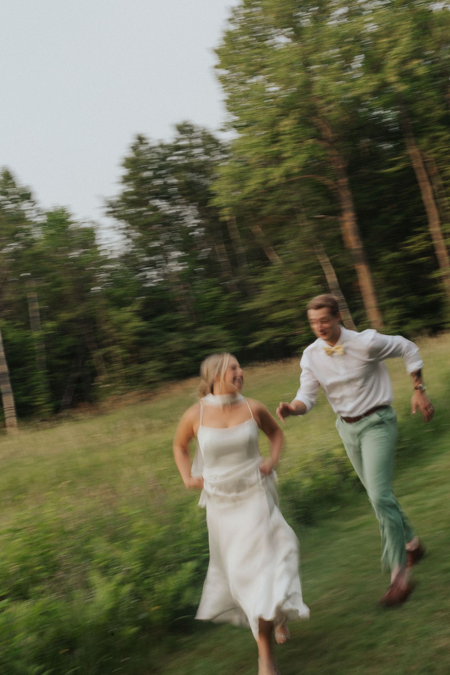 Joyful bride and groom run through a green meadow with a blurred motion effect near a forest.