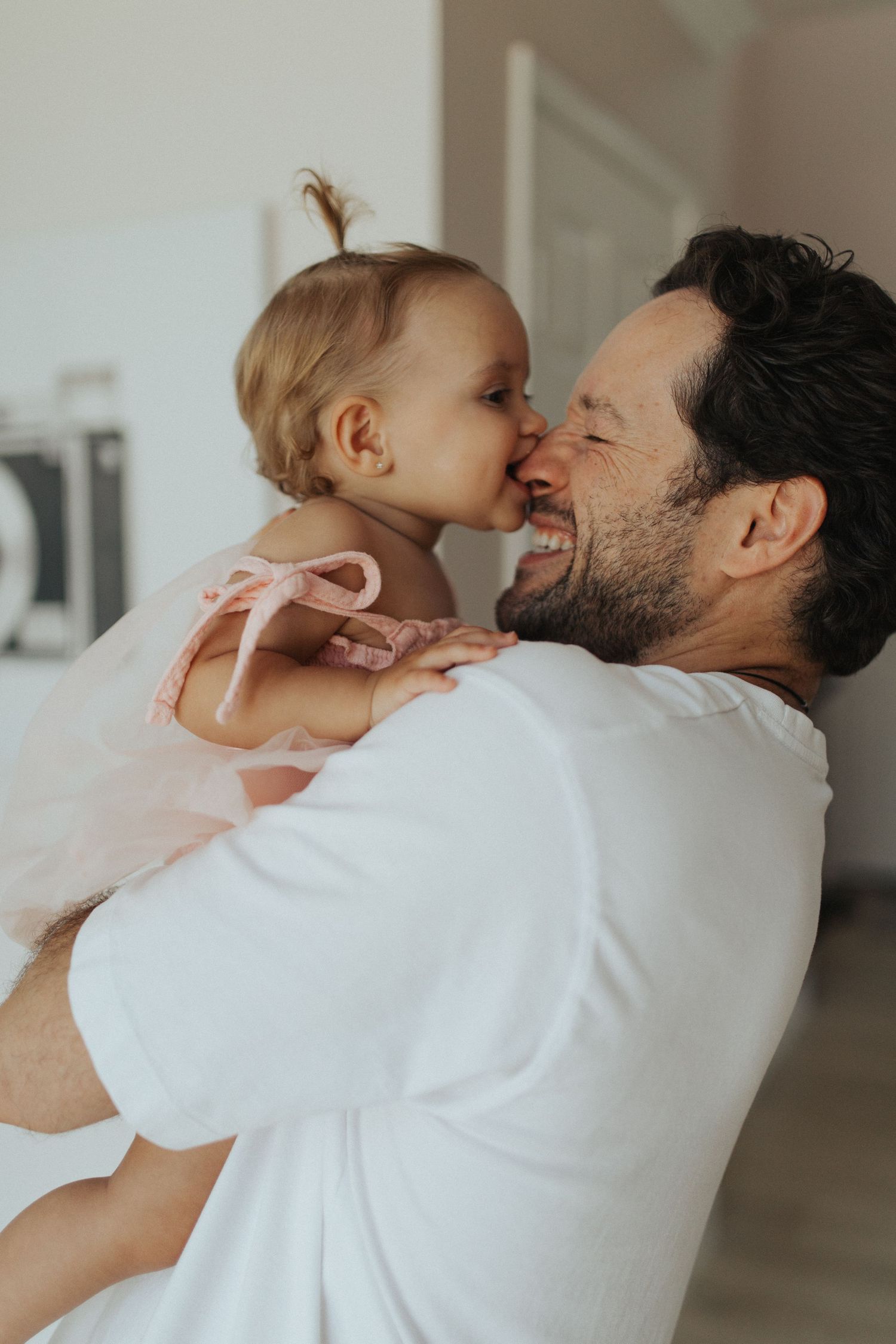 Father holds baby girl in pink dress as she kisses his cheek.