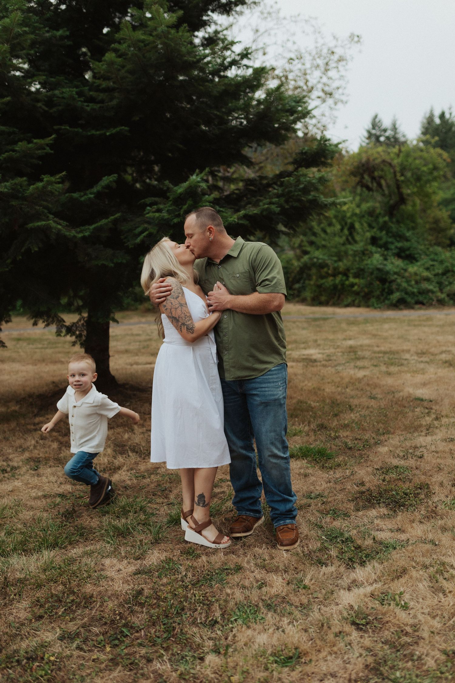 Couple sharing a kiss outdoors while their young son runs playfully beside them in a park.