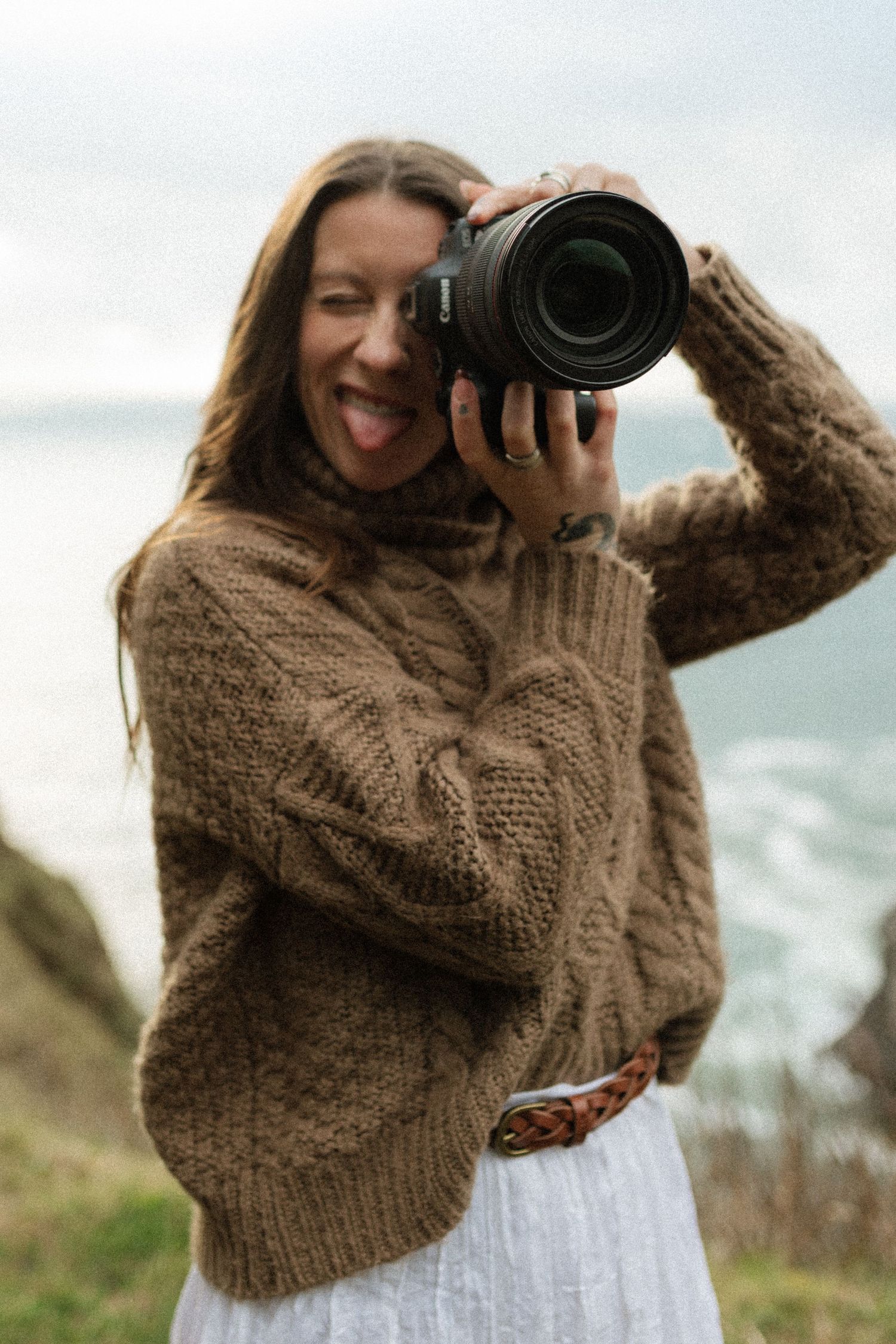 Playful female photographer in brown cable-knit sweater holds camera up with tongue out near a Oregon coastal cliffside.