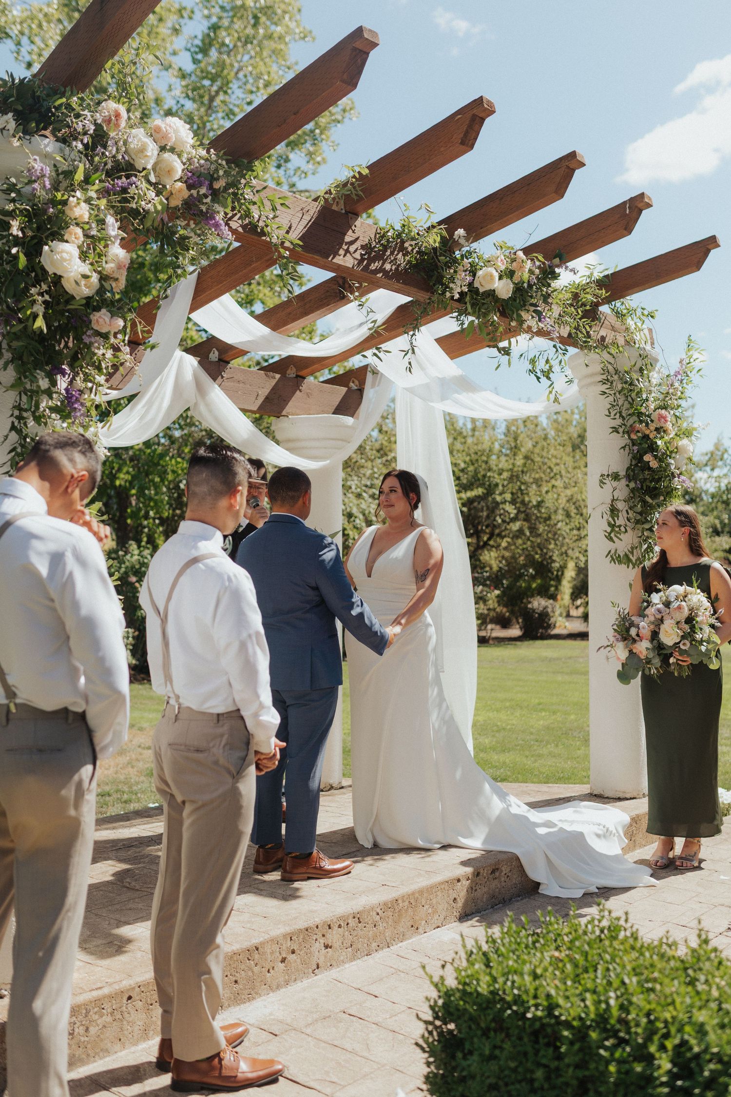 Bride and groom exchange vows under a floral-adorned wooden pergola at a sunny outdoor garden wedding ceremony.