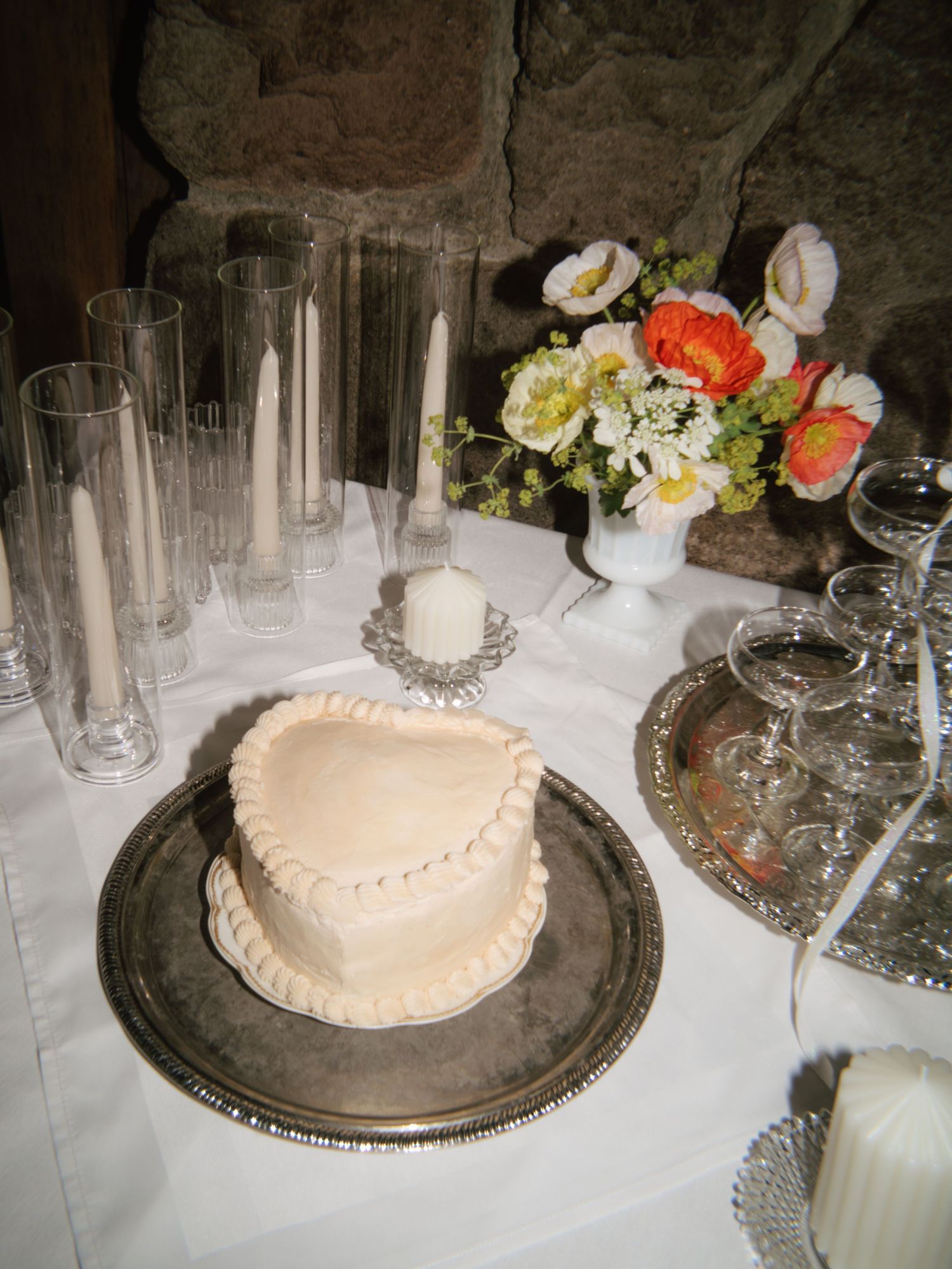 Heart-shaped white wedding cake on silver tray with flowers and candles on table.