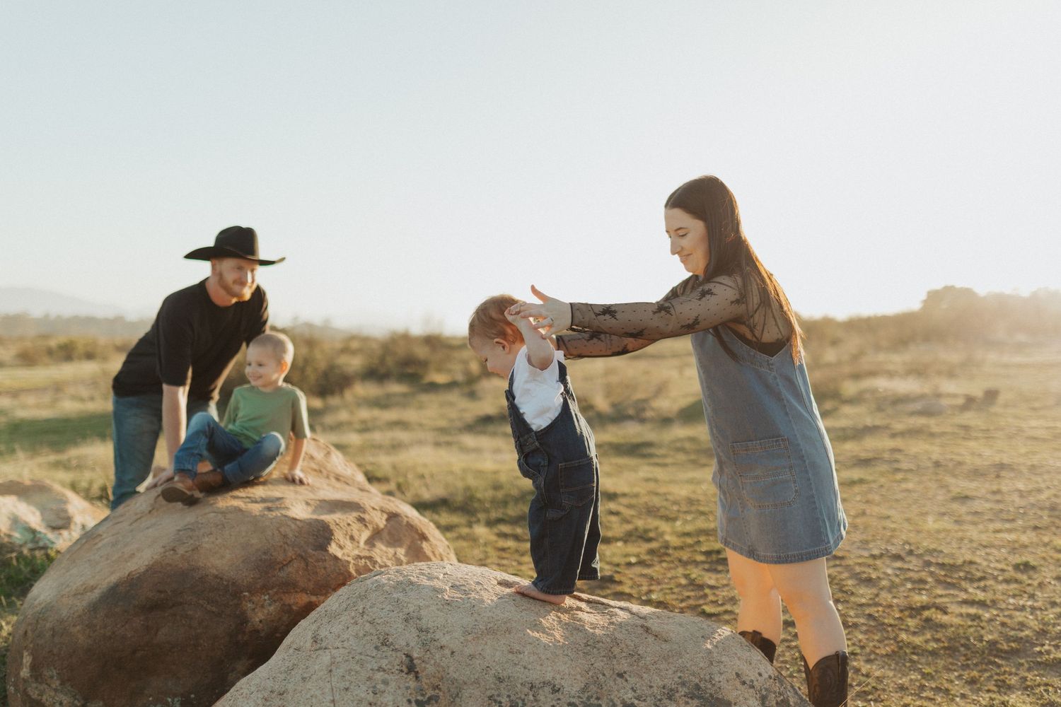 Two women helping young children climb large boulders in a sunny open field during family photo session.