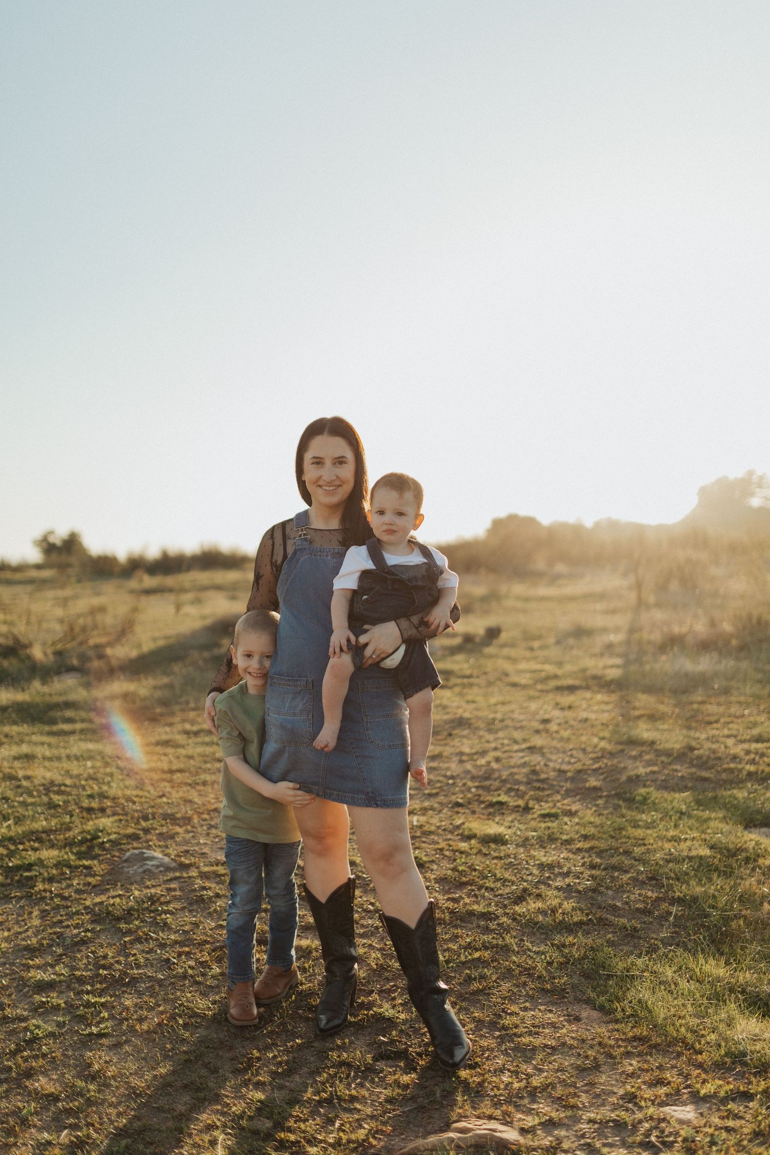 Mother in denim dress and boots holds baby in carrier while young son hugs her in sunny field.