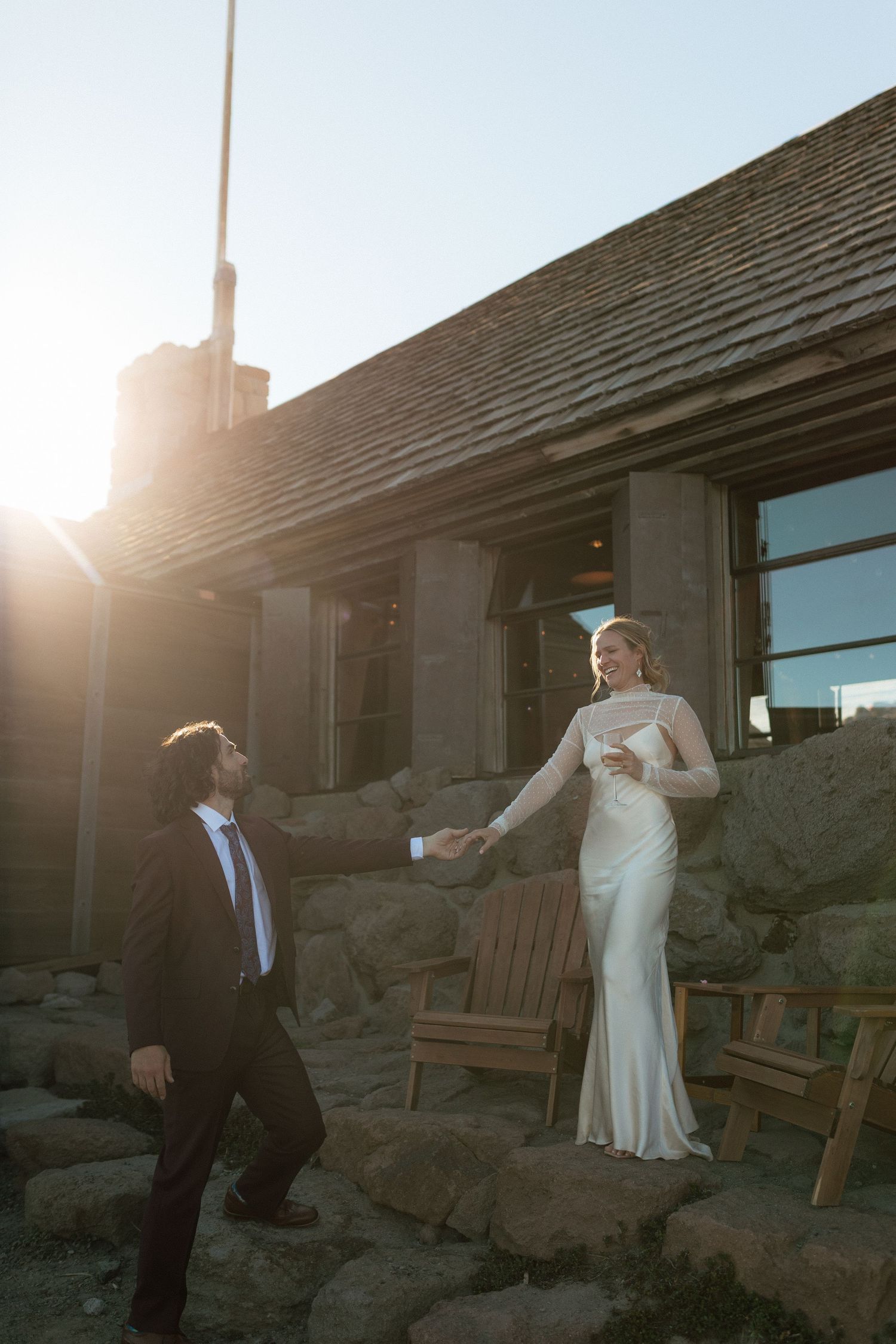 Bride and groom hold hands at golden hour outside a rustic stone lodge wedding venue.