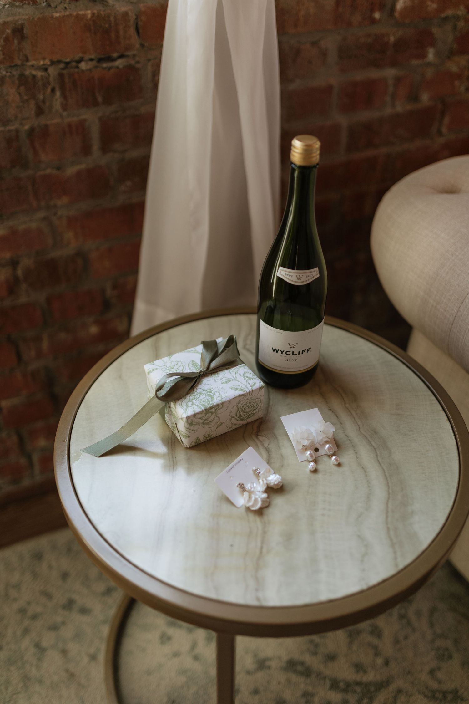 Wedding details flat lay with champagne bottle, pearl earrings, and gift box on marble table.