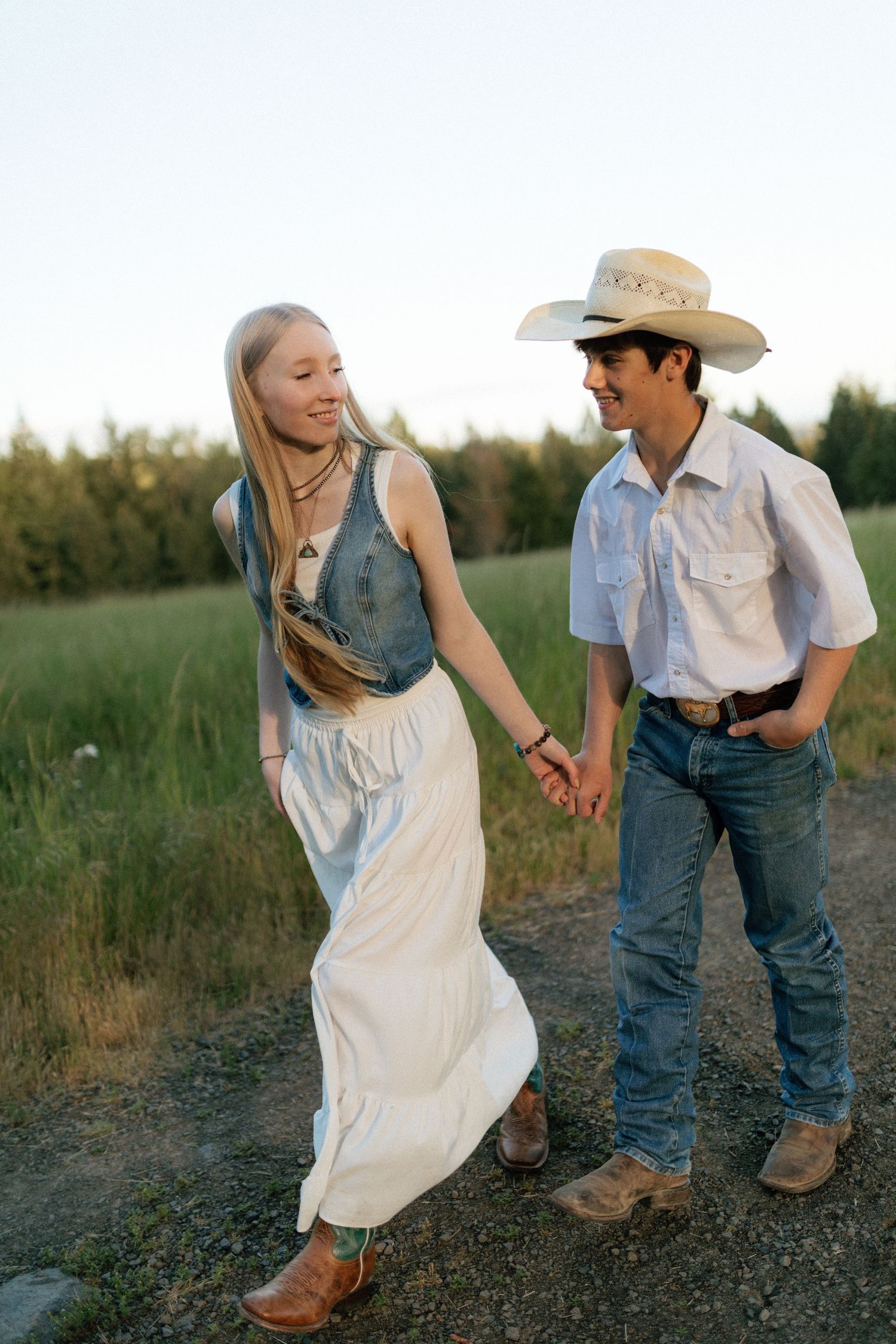 Smiling couple walking hand in hand on a gravel path, woman in white skirt and denim vest.