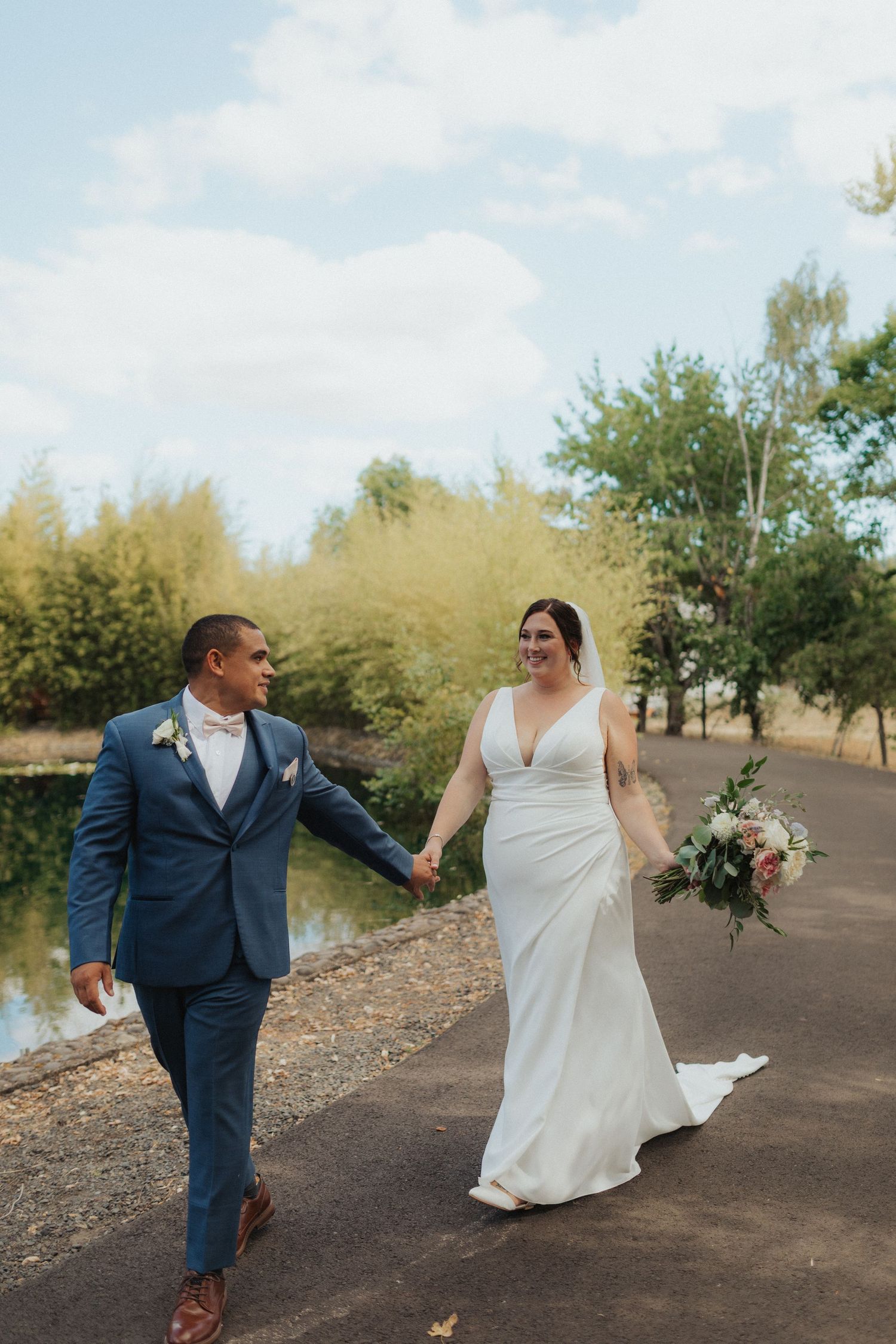 Bride and groom walk hand-in-hand along a pond path, bride holding a pink bouquet on their wedding day.