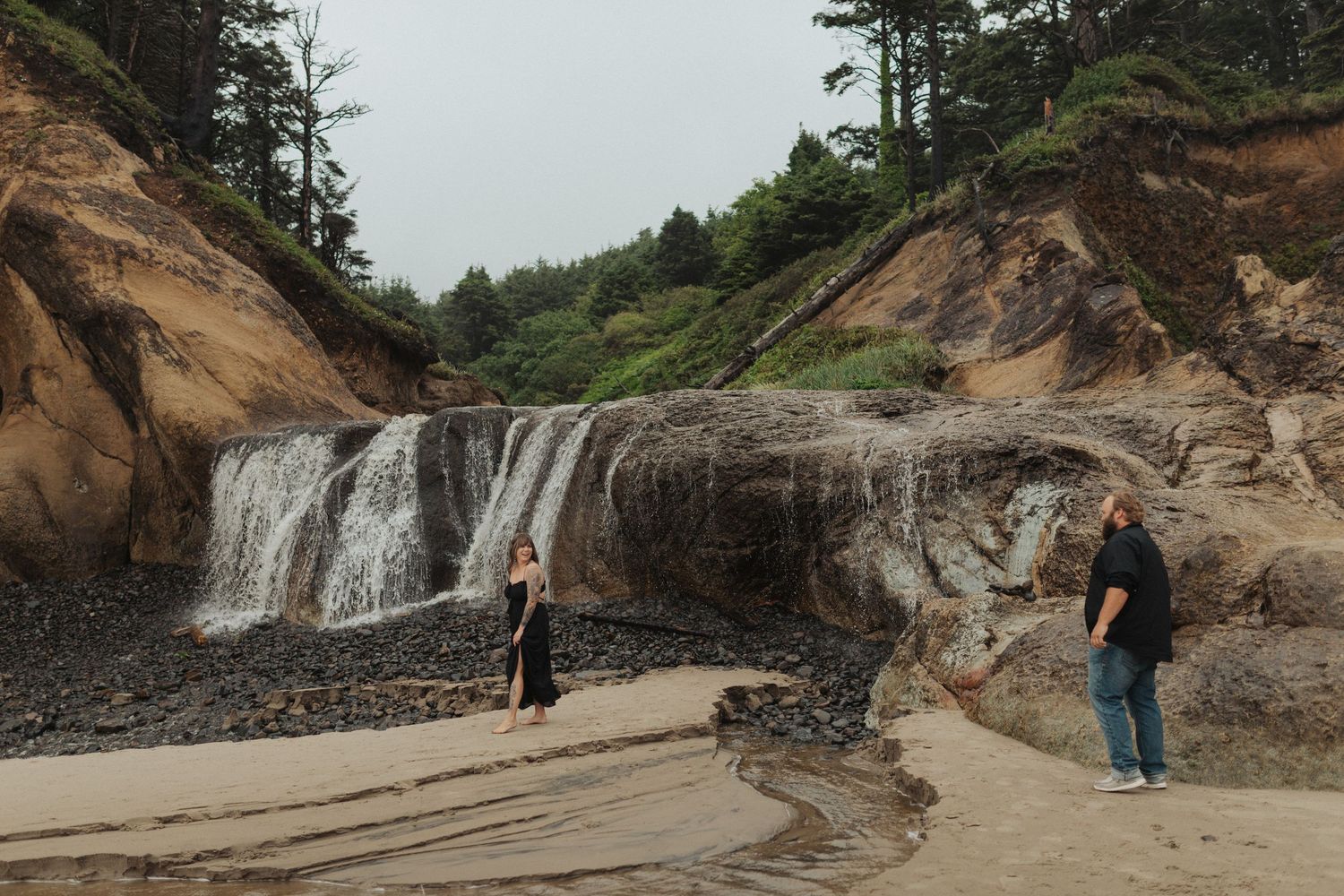 Couple exploring a scenic beach with a cascading waterfall and sandy cliffs surrounded by lush trees.