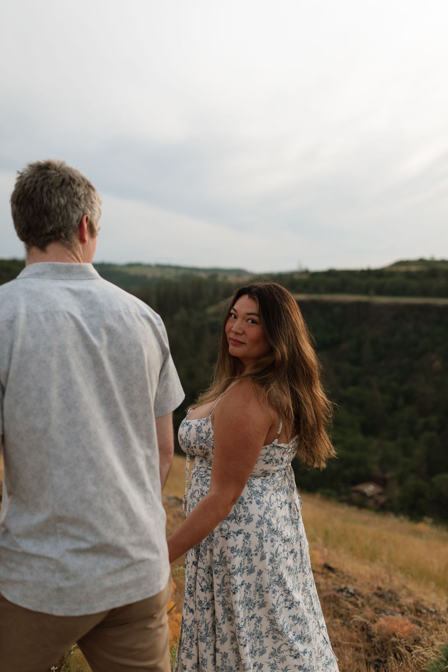 Woman in floral dress looks back smiling while holding hands with man on scenic hilltop.