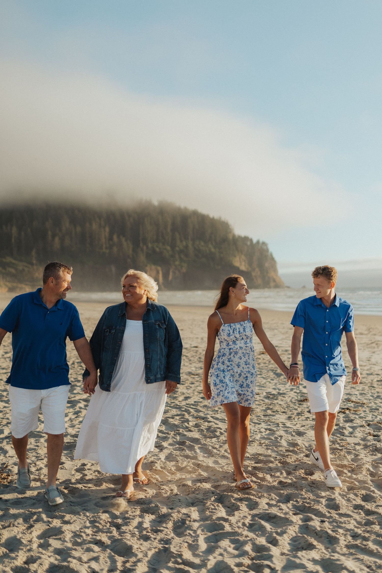 A family of four walks hand in hand along a sandy beach with misty coastal cliffs in the background.