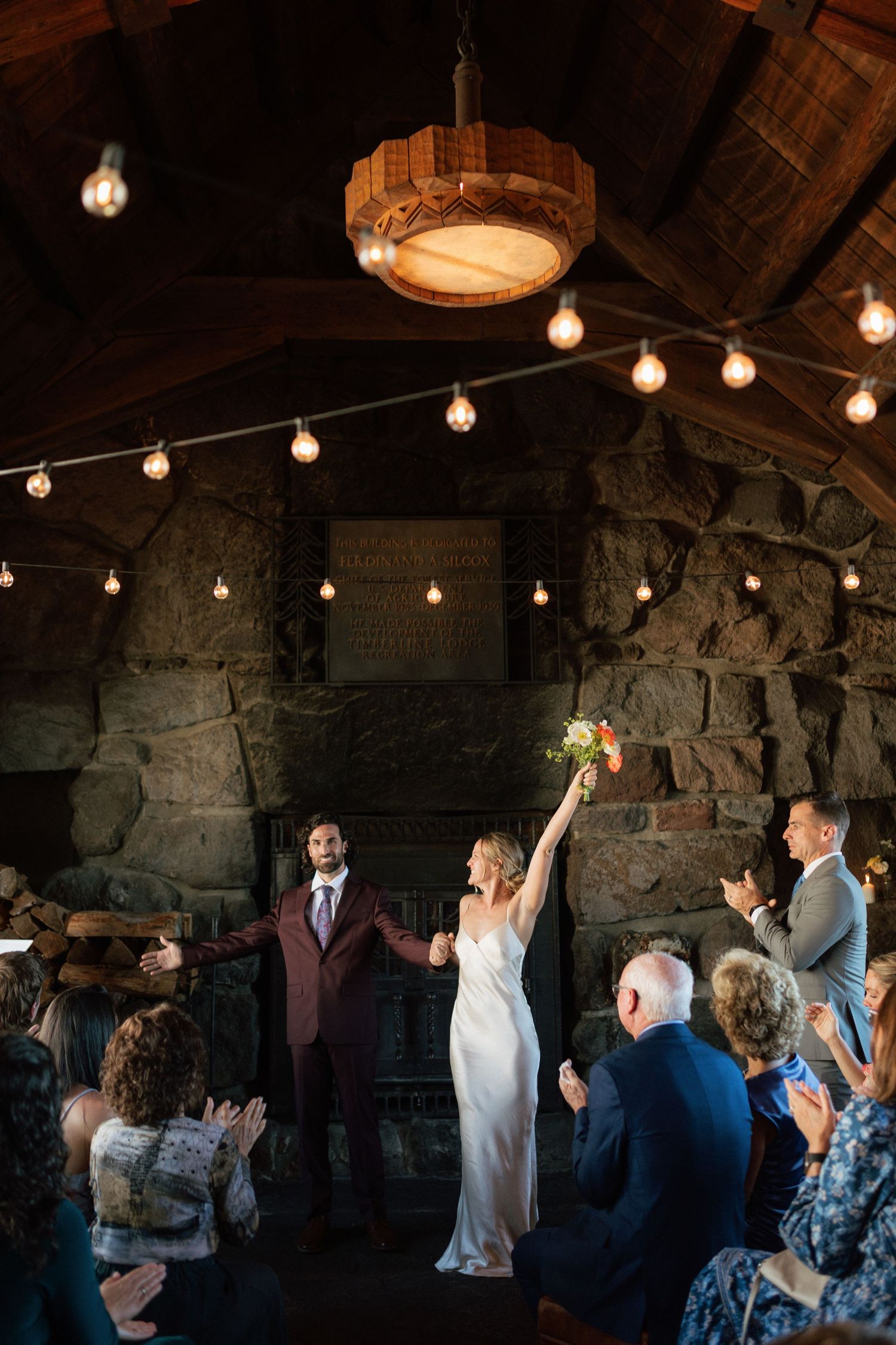 Newlyweds celebrate with raised hands at rustic stone venue adorned with string lights during wedding ceremony.