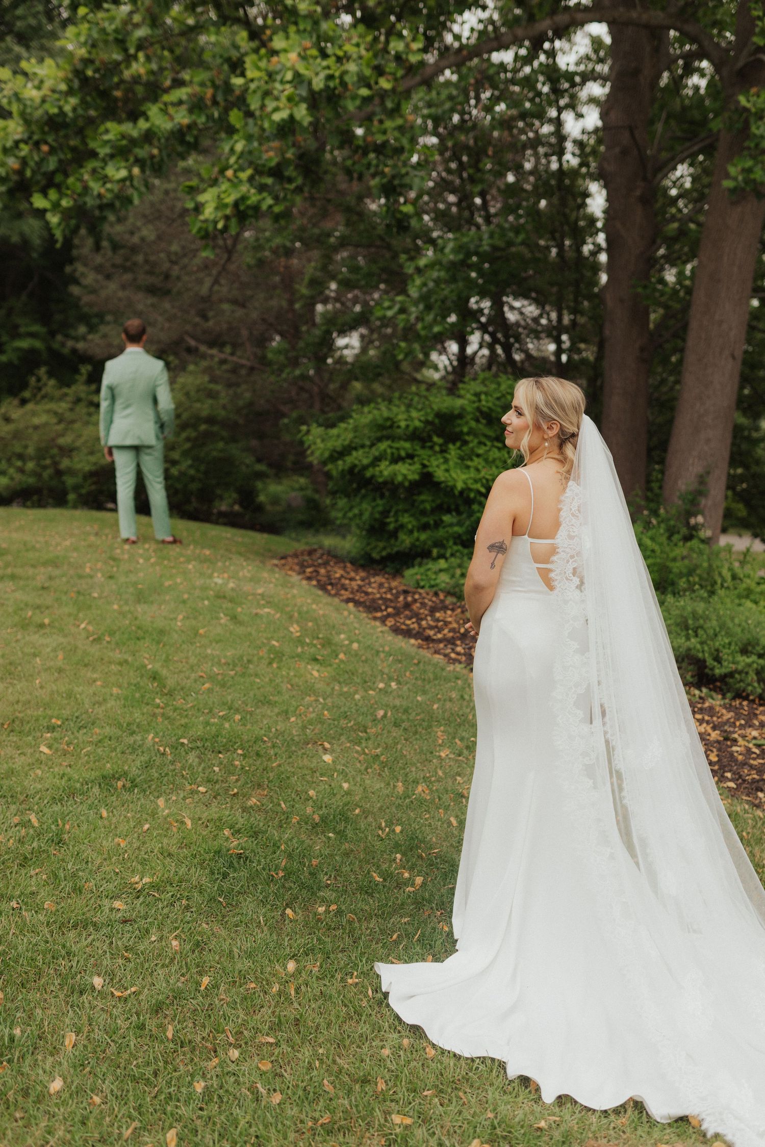 Bride in a white gown with long veil walks toward groom in mint suit during outdoor first look.