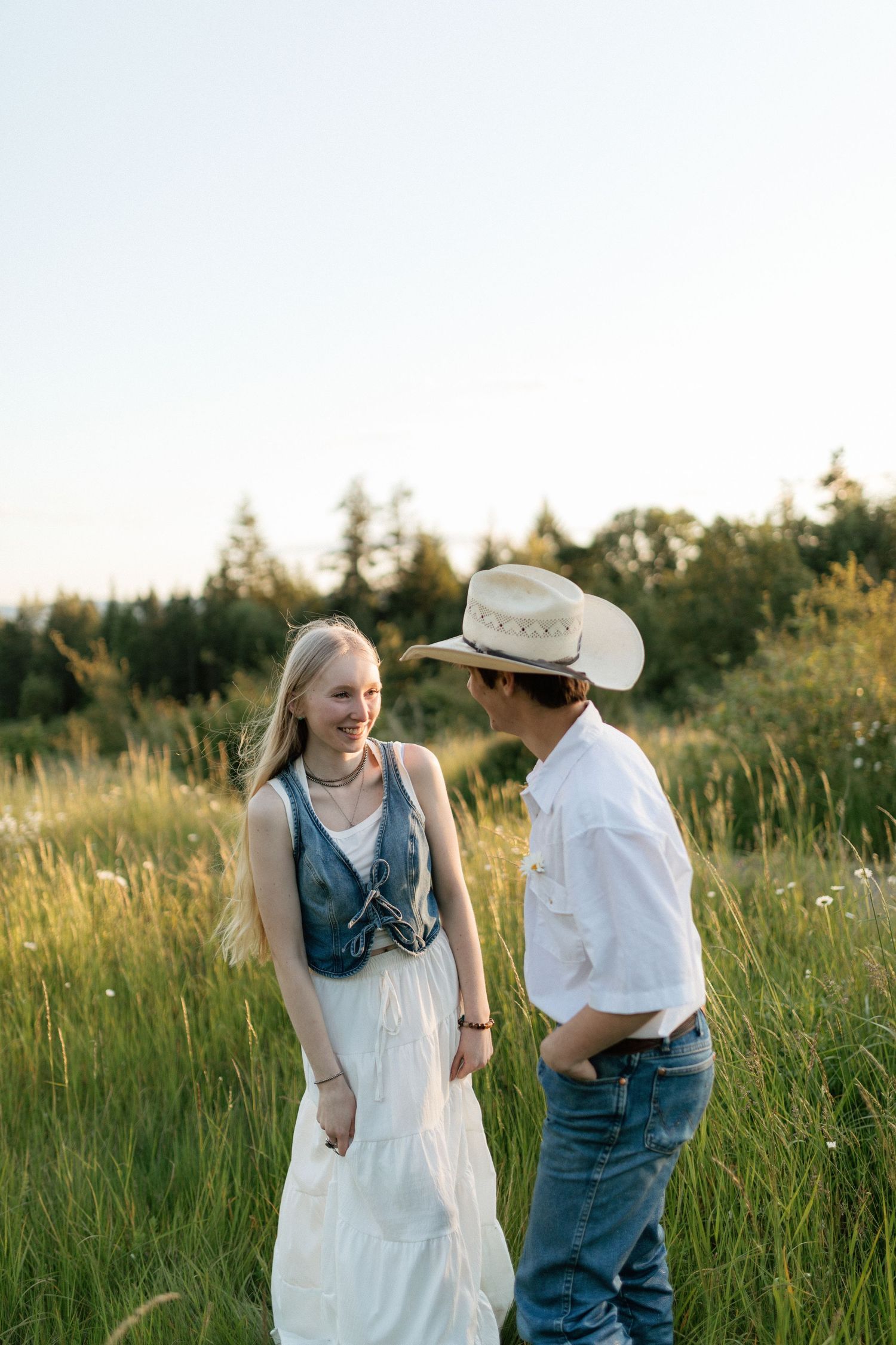 Smiling young couple standing in sunlit meadow, woman in white dress, man in cowboy hat and jeans.