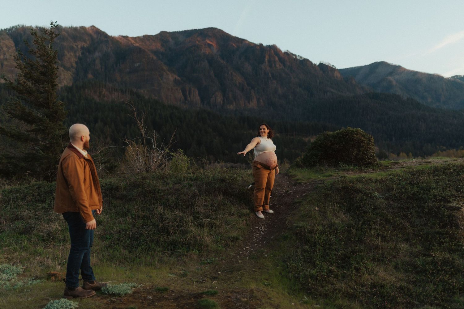 Couple standing apart on a mountain trail at dusk with evergreen trees and rocky peaks in the background.