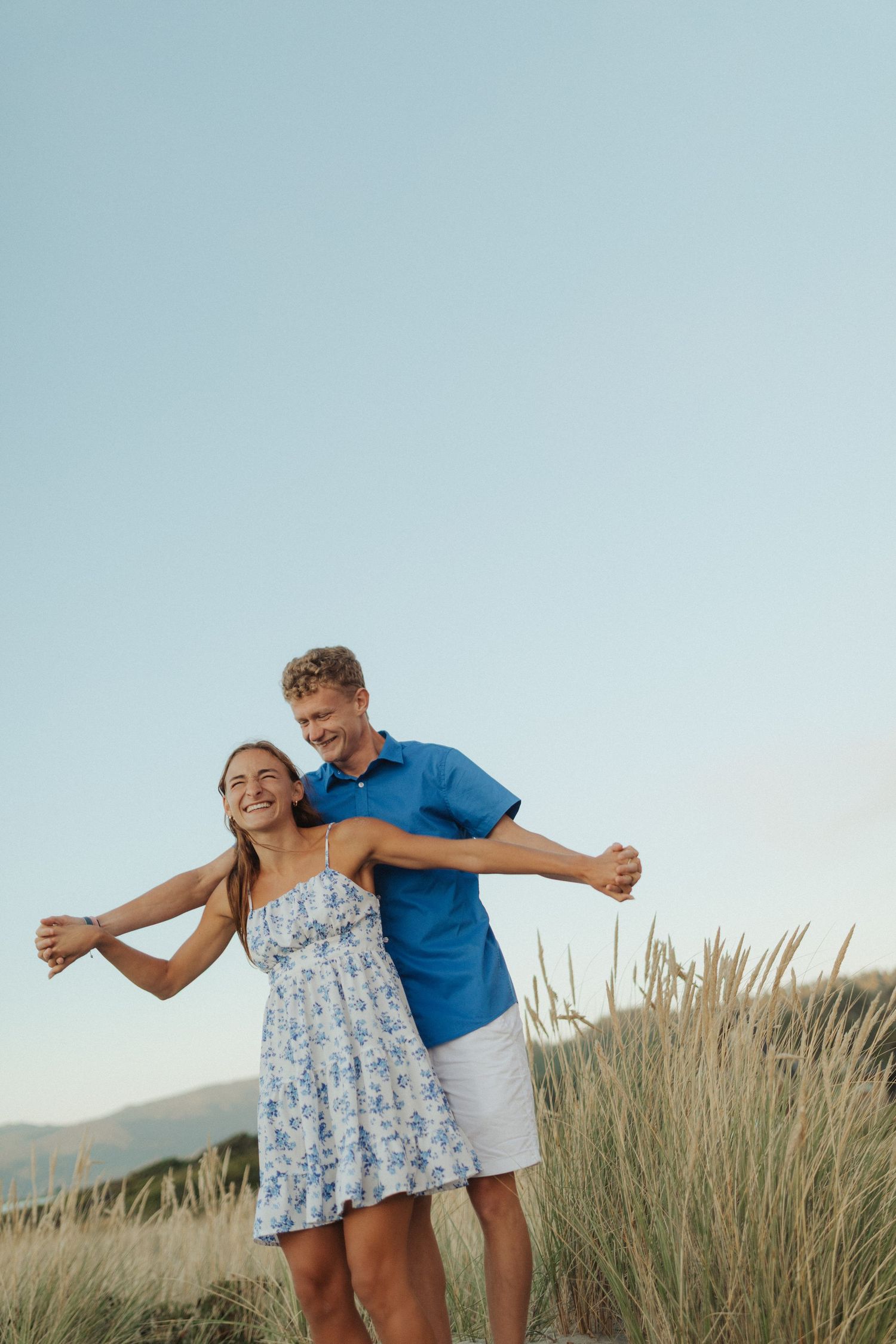 Happy young couple embracing with arms outstretched in tall beach grass under a clear blue sky.