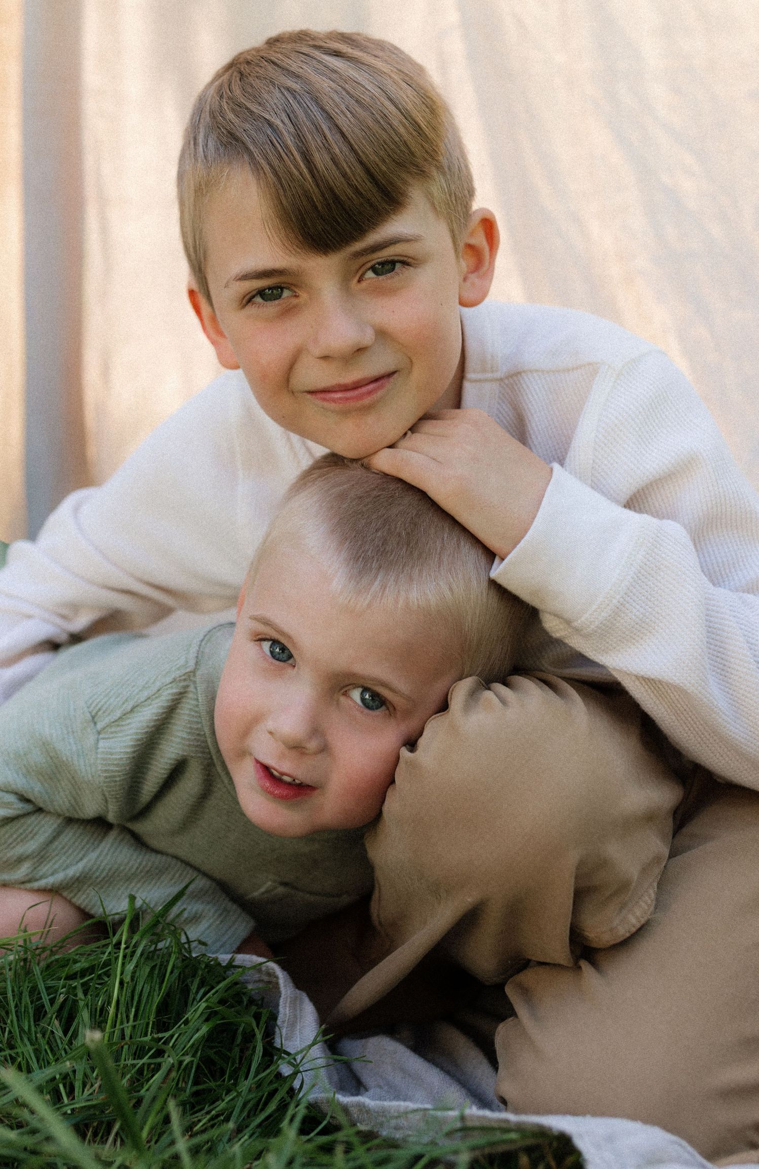 Two brothers cuddle together on grass in front of a neutral fabric backdrop in a tender portrait.