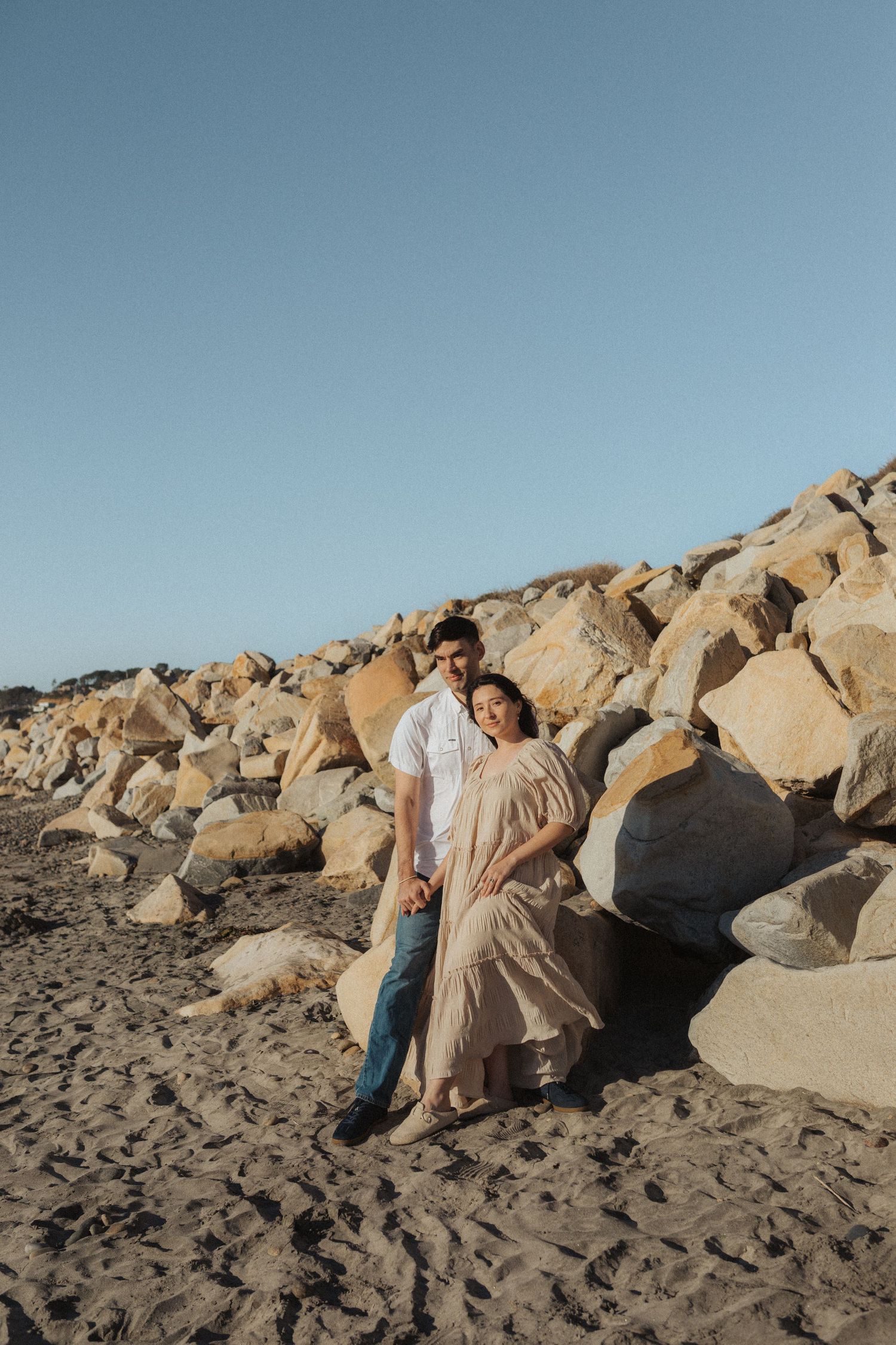Couple posing together against large boulders on a sandy beach, woman in elegant flowing beige maxi dress.