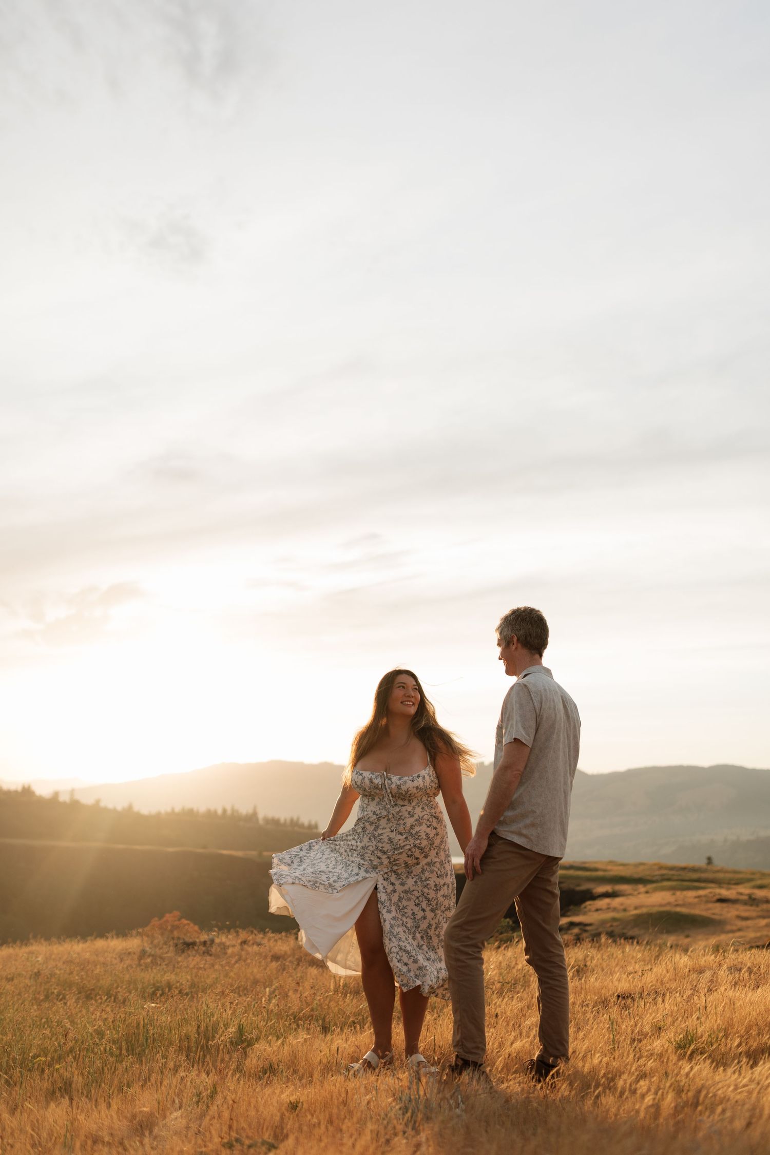Couple holding hands in a golden field at sunset with warm light glowing behind the hills.