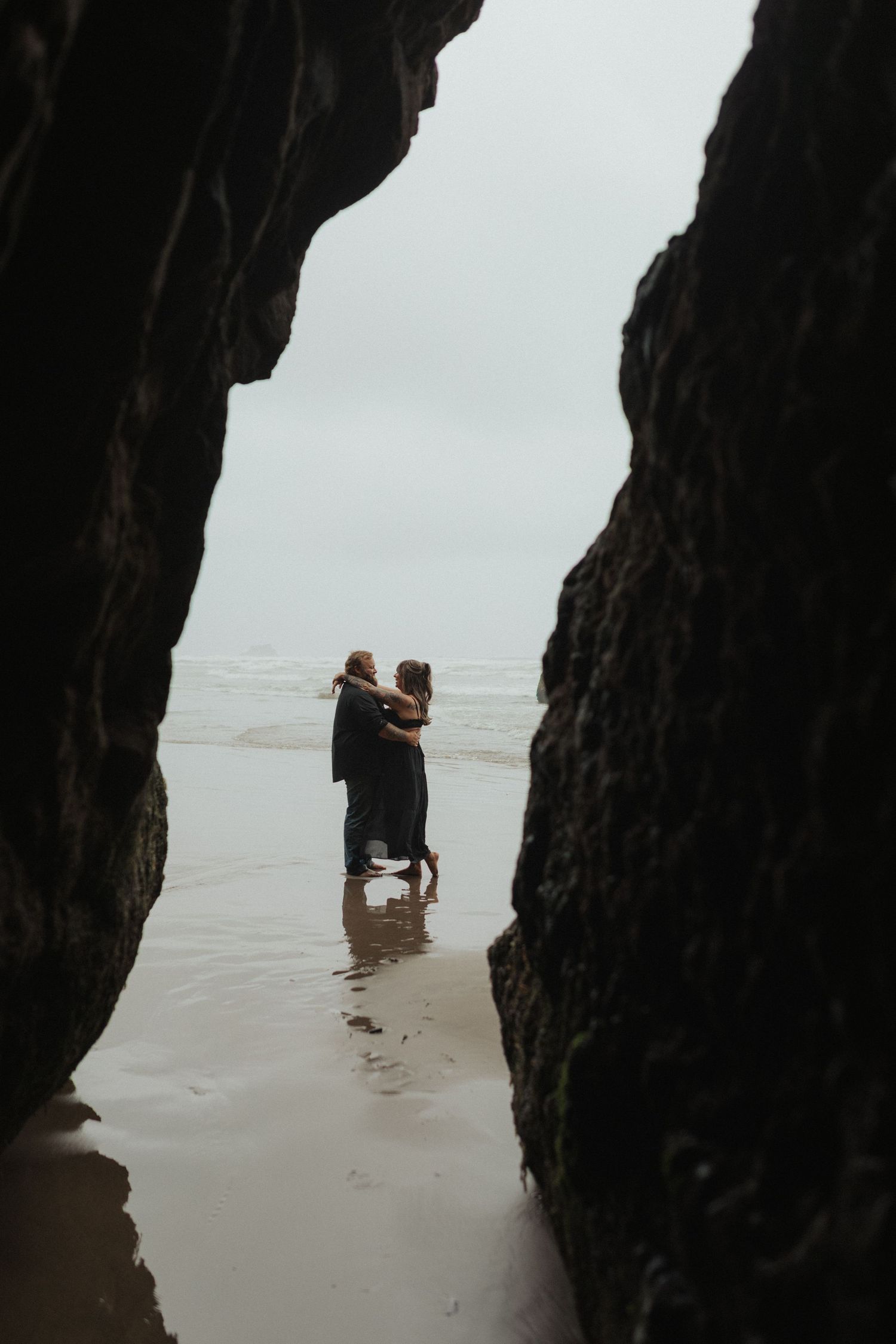 Couple embracing romantically between dark rock formations on a misty beach shoreline.