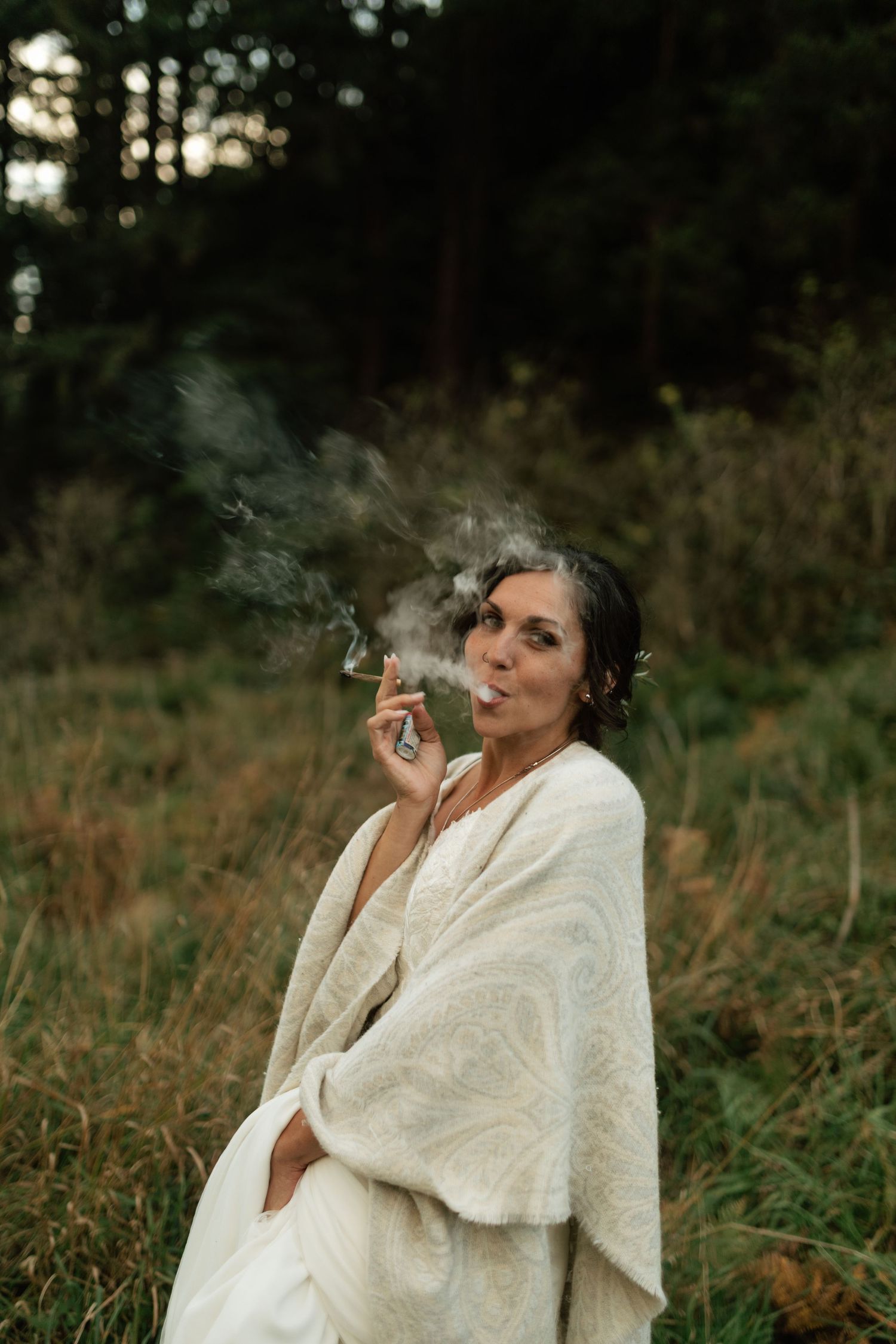 Bride in white shawl smoking outdoors in a meadow with forest background.