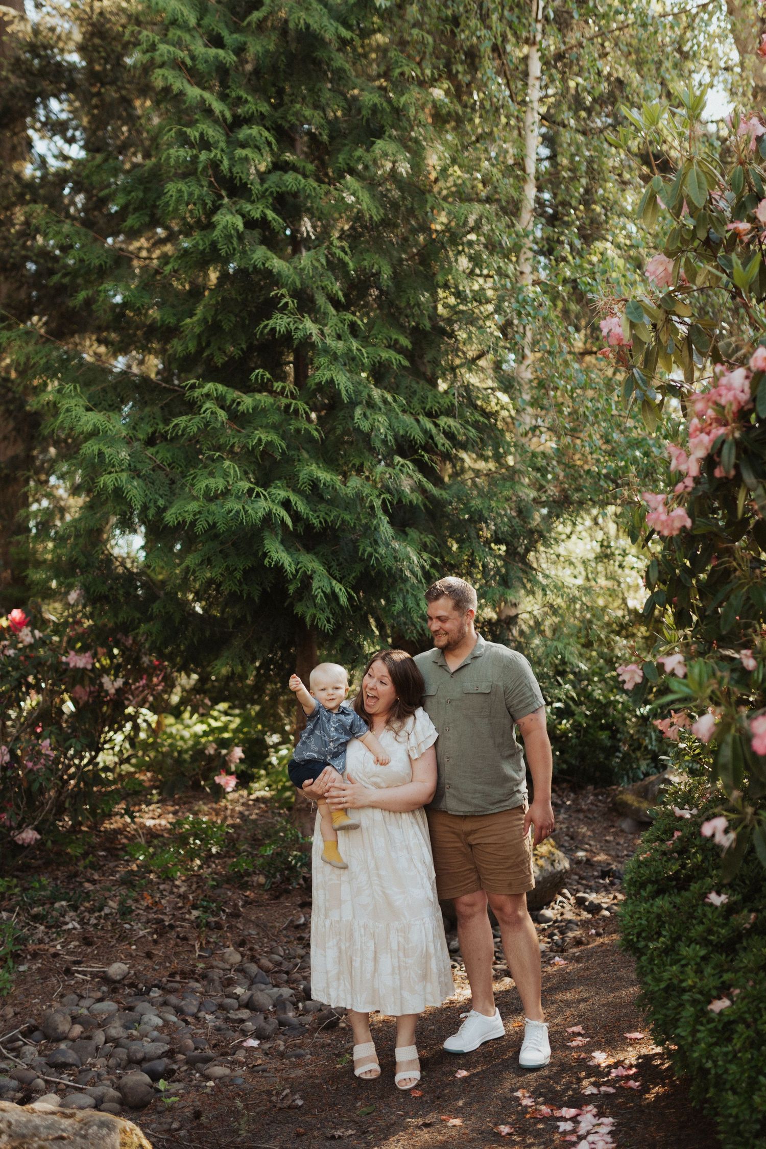 A happy family with a baby walks along a garden path surrounded by lush green trees and pink flowers.