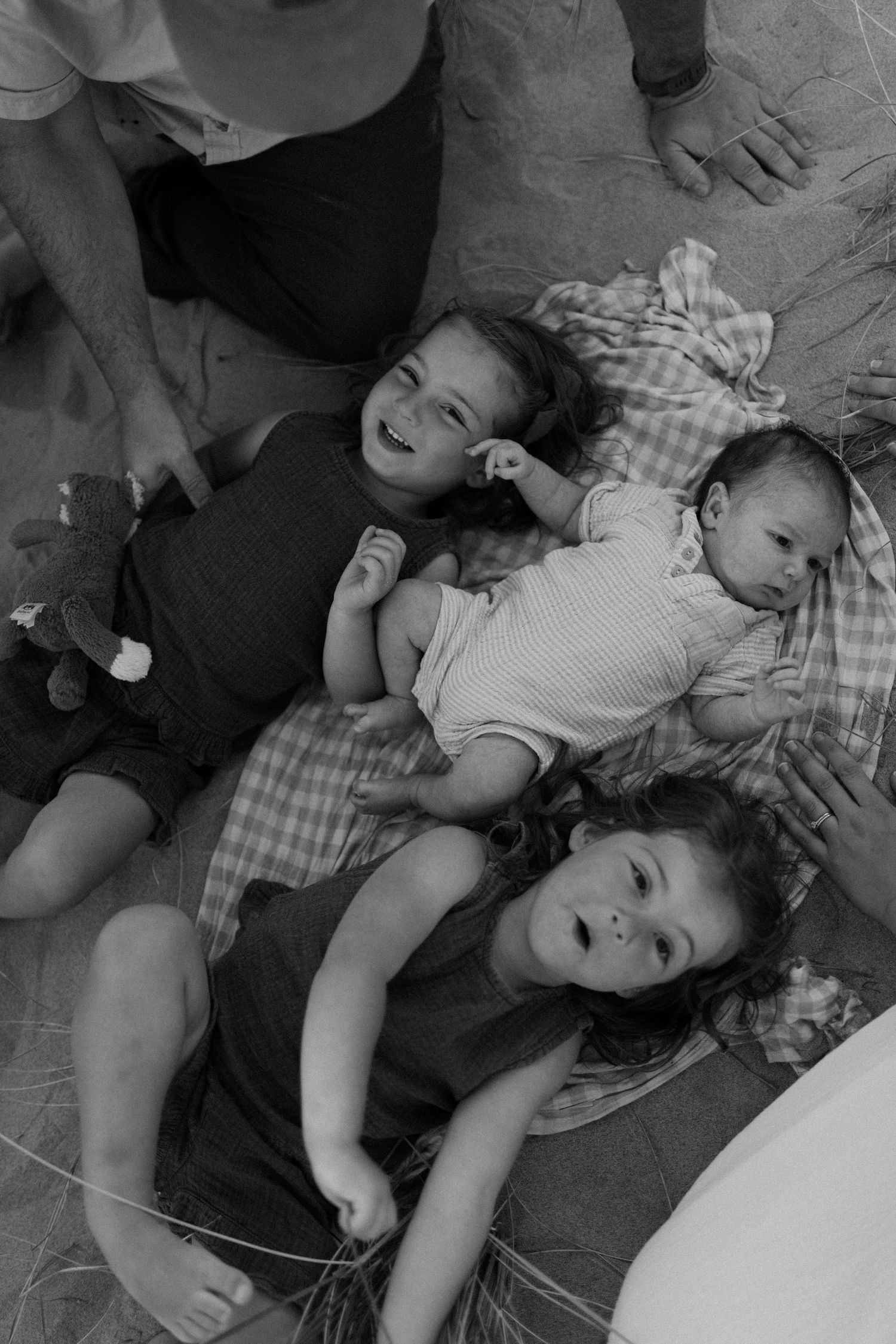 Three children lying on a blanket in the sand, including a newborn baby, captured in black and white.