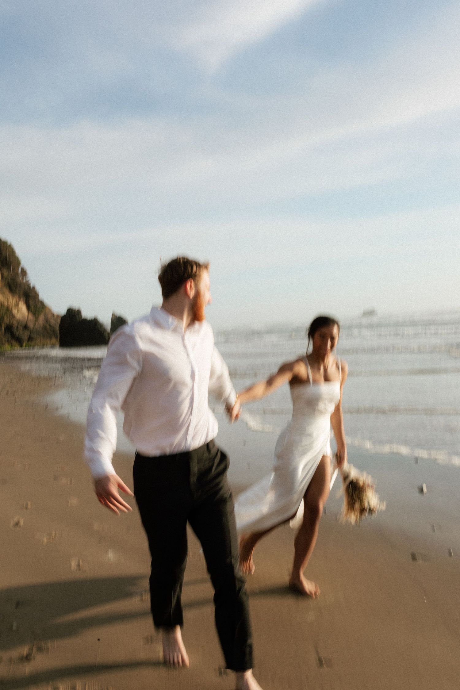 Motion-blurred photo of barefoot bride and groom running playfully along ocean shoreline during golden hour.