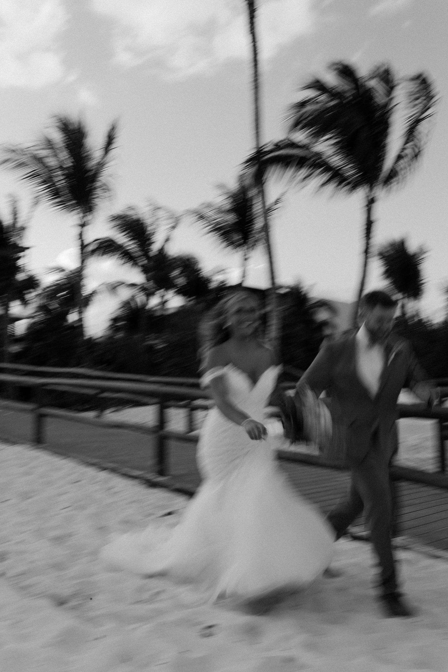 Bride and groom run joyfully along a sandy beach at sunset in a blurred black and white wedding photo.