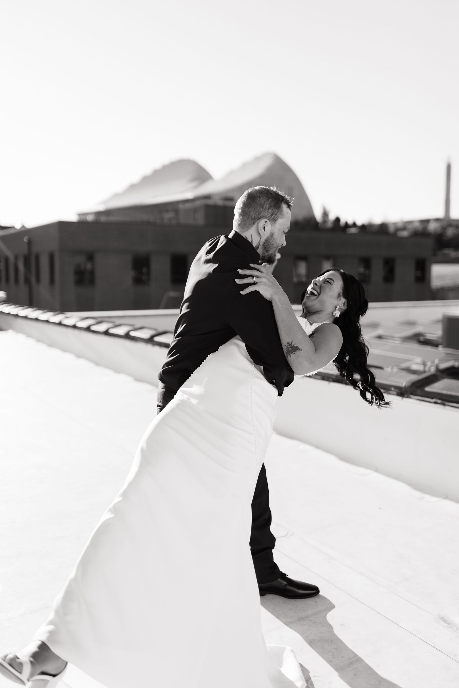 Black and white photo of a bride and groom sharing a joyful moment on a rooftop, with the bride laughing in her gown.