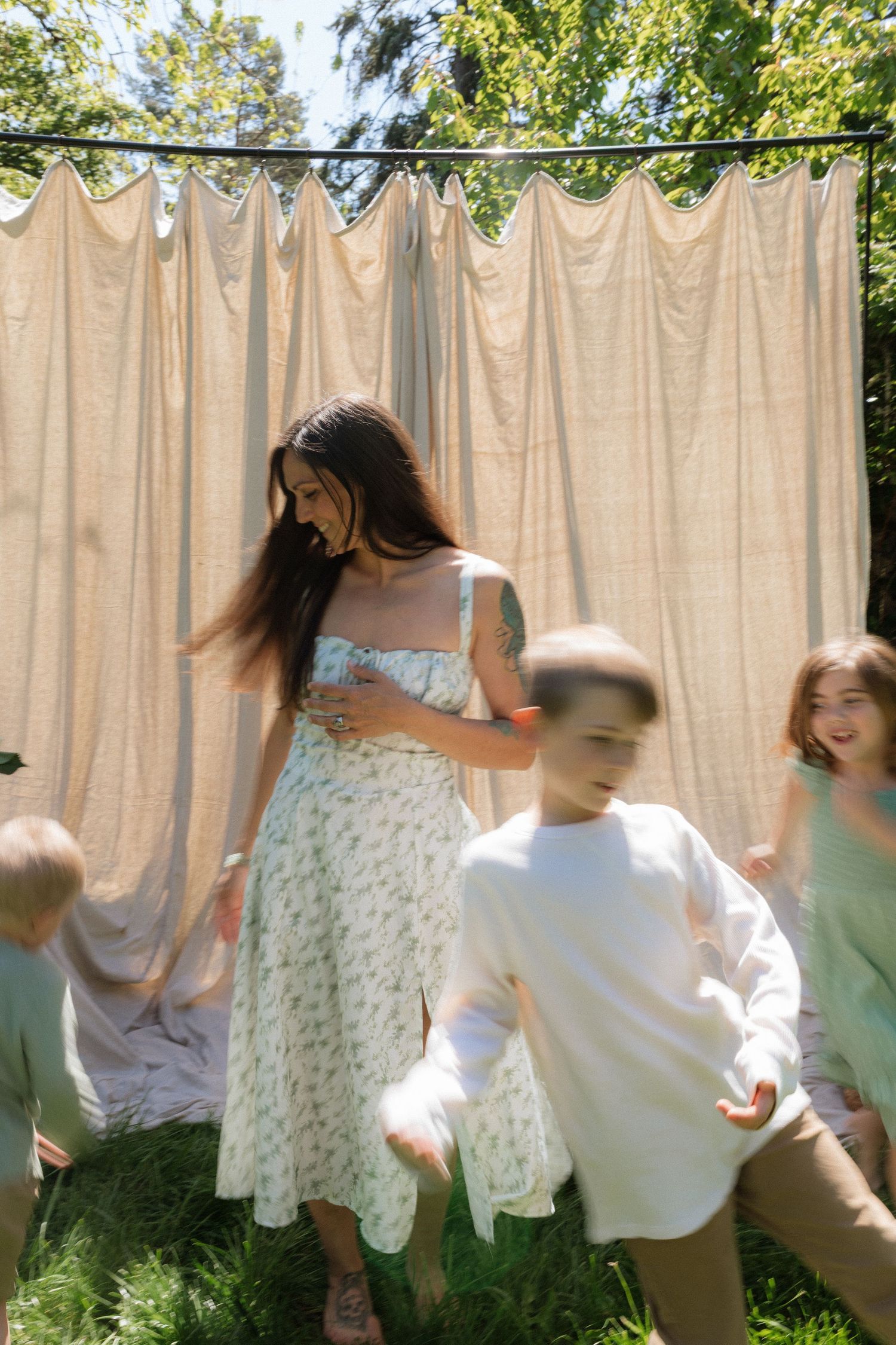A smiling woman in a floral dress plays with energetic children in front of a beige curtain outdoors.