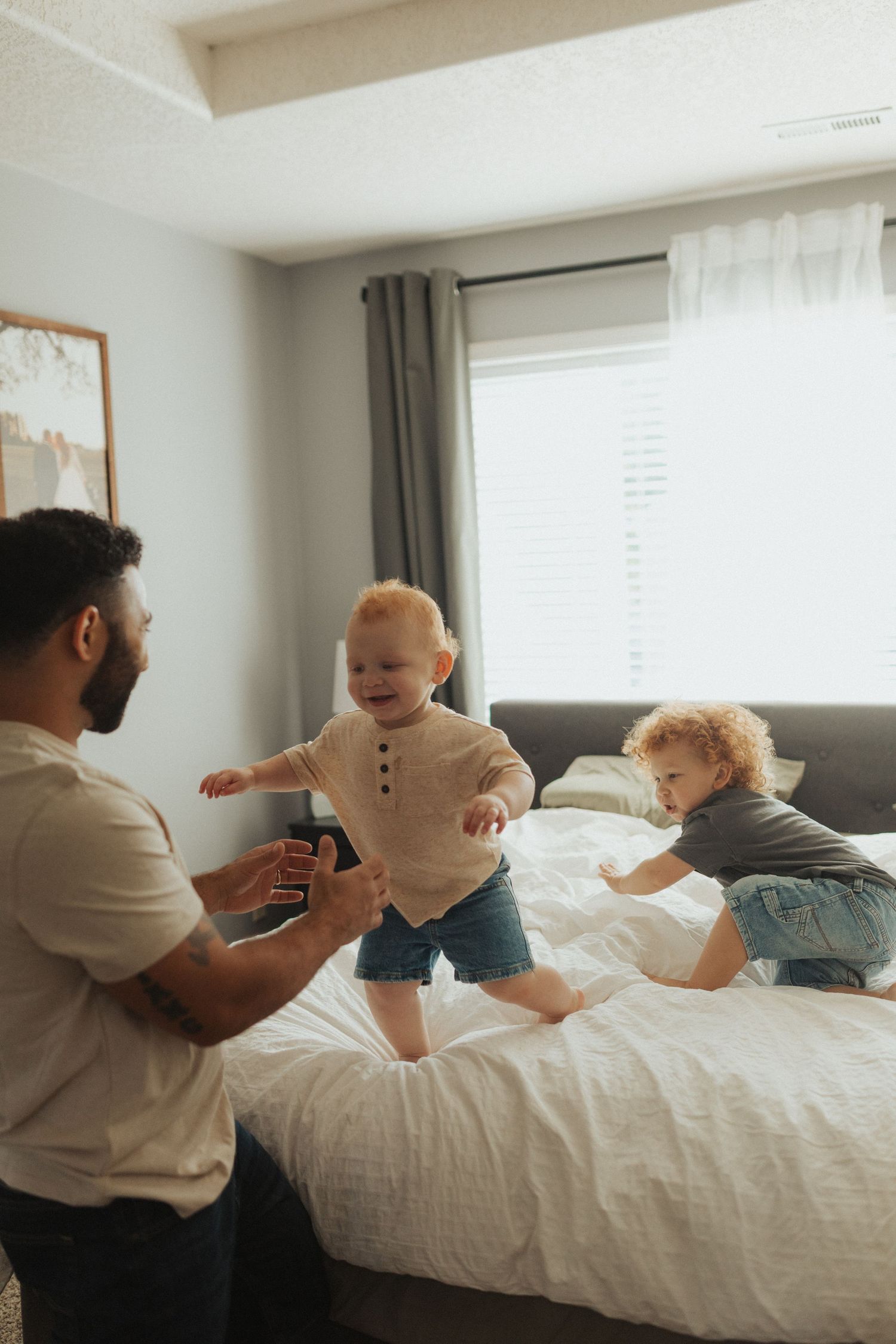 A father plays with two young toddlers jumping and crawling on a white bed in a cozy bedroom.