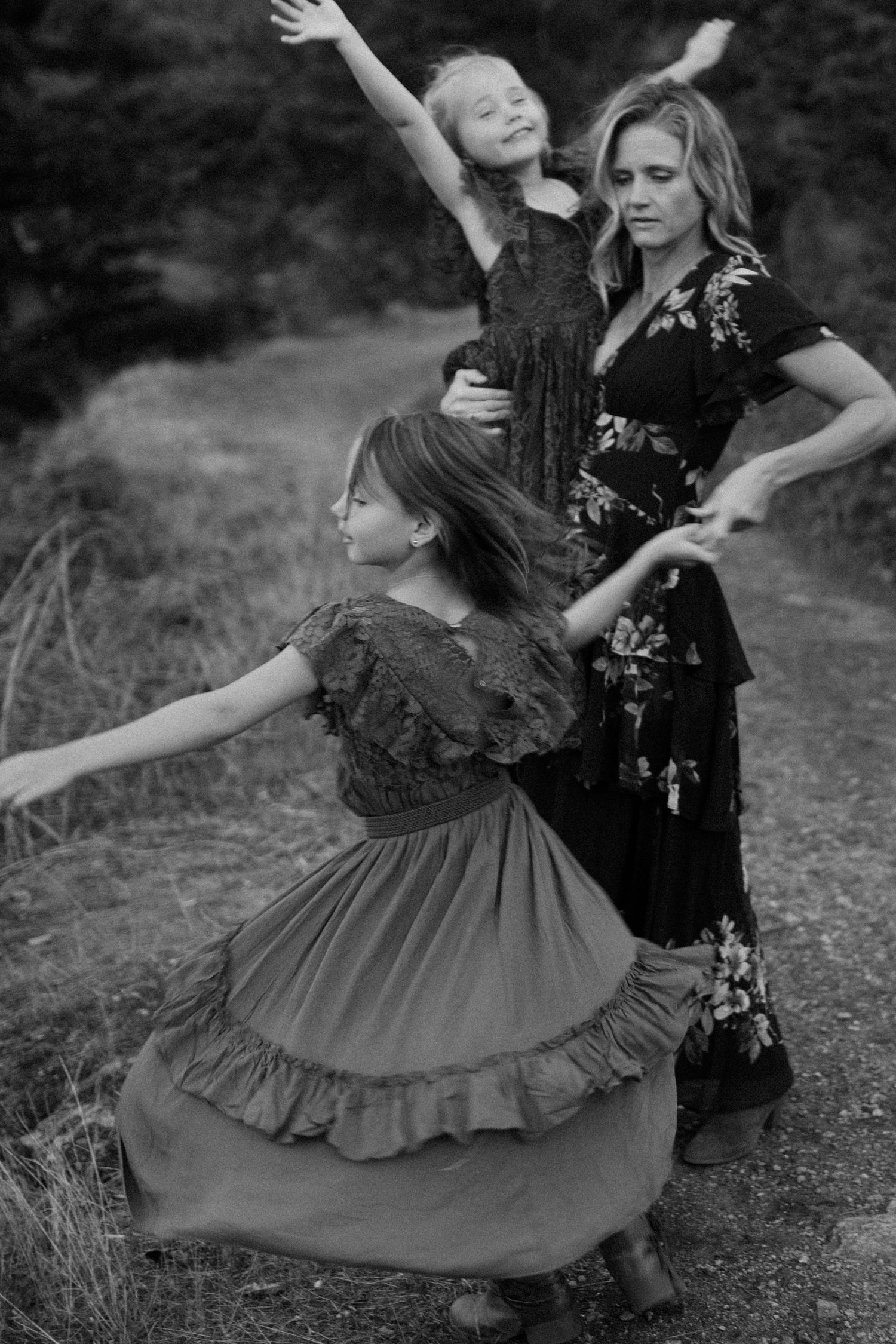 Black and white photo of a mother and two daughters dancing freely on a forest path together.