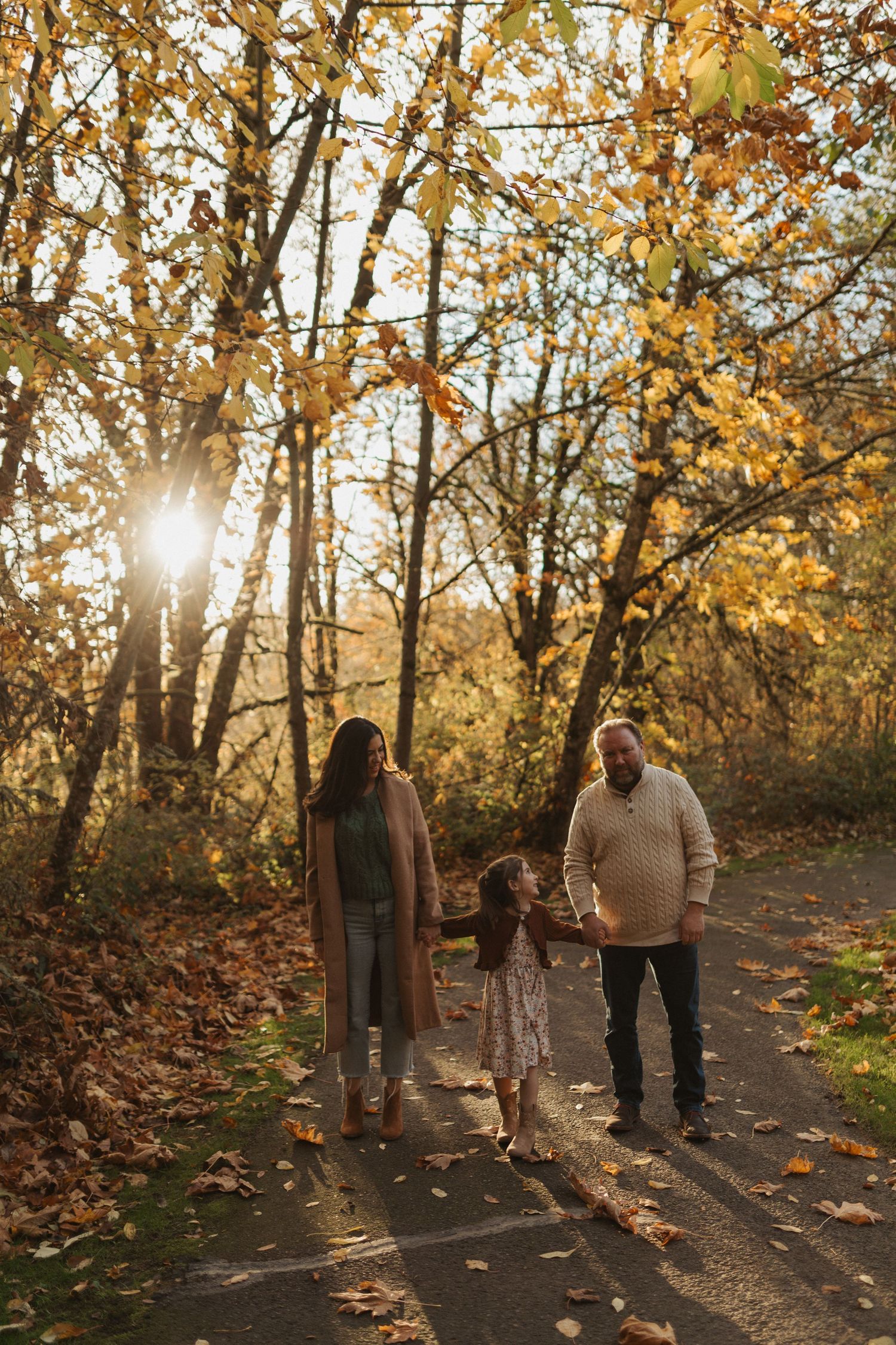 Family of three walking hand in hand on a sunlit autumn trail with golden leaves and warm sunlight.