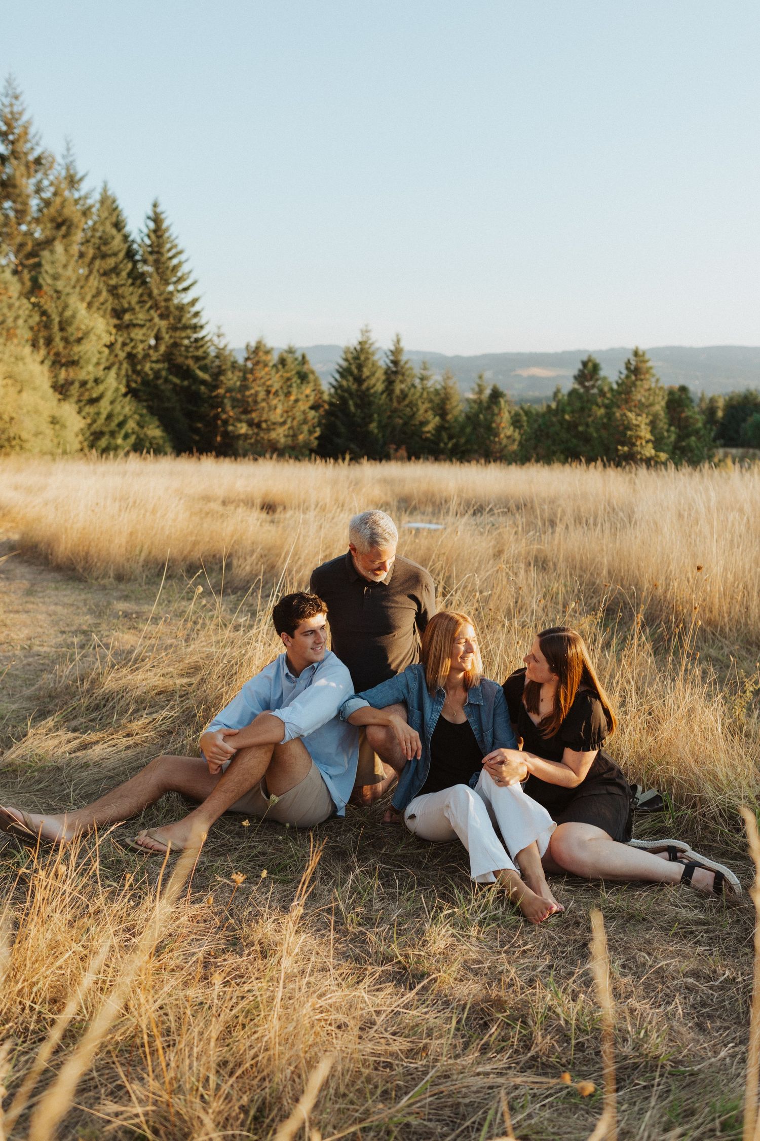Family of four sitting together in a golden dry grass field surrounded by evergreen trees.
