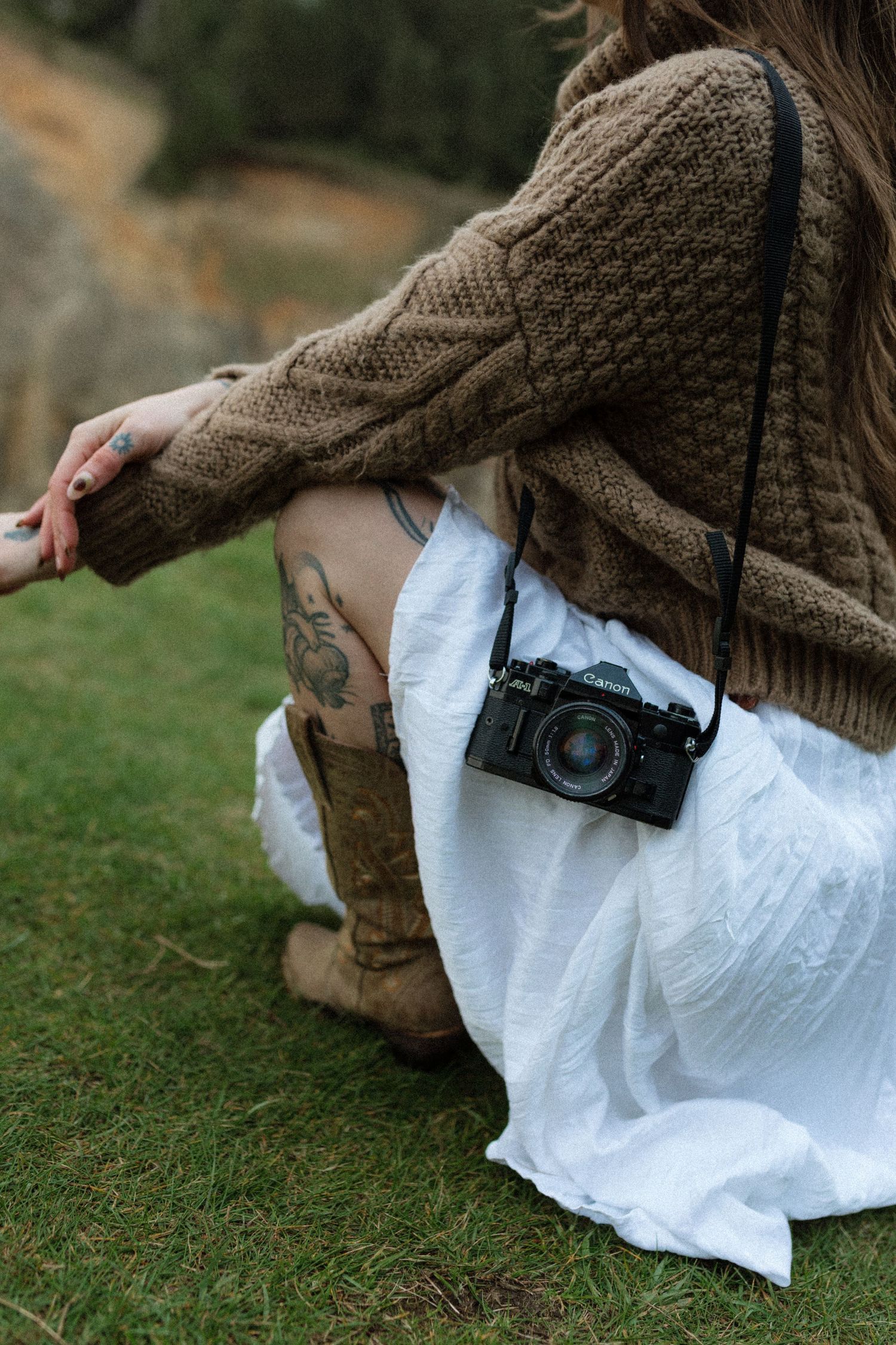 Close-up of a woman in a brown knit sweater with leg tattoos, holding a vintage Canon film camera outdoors.