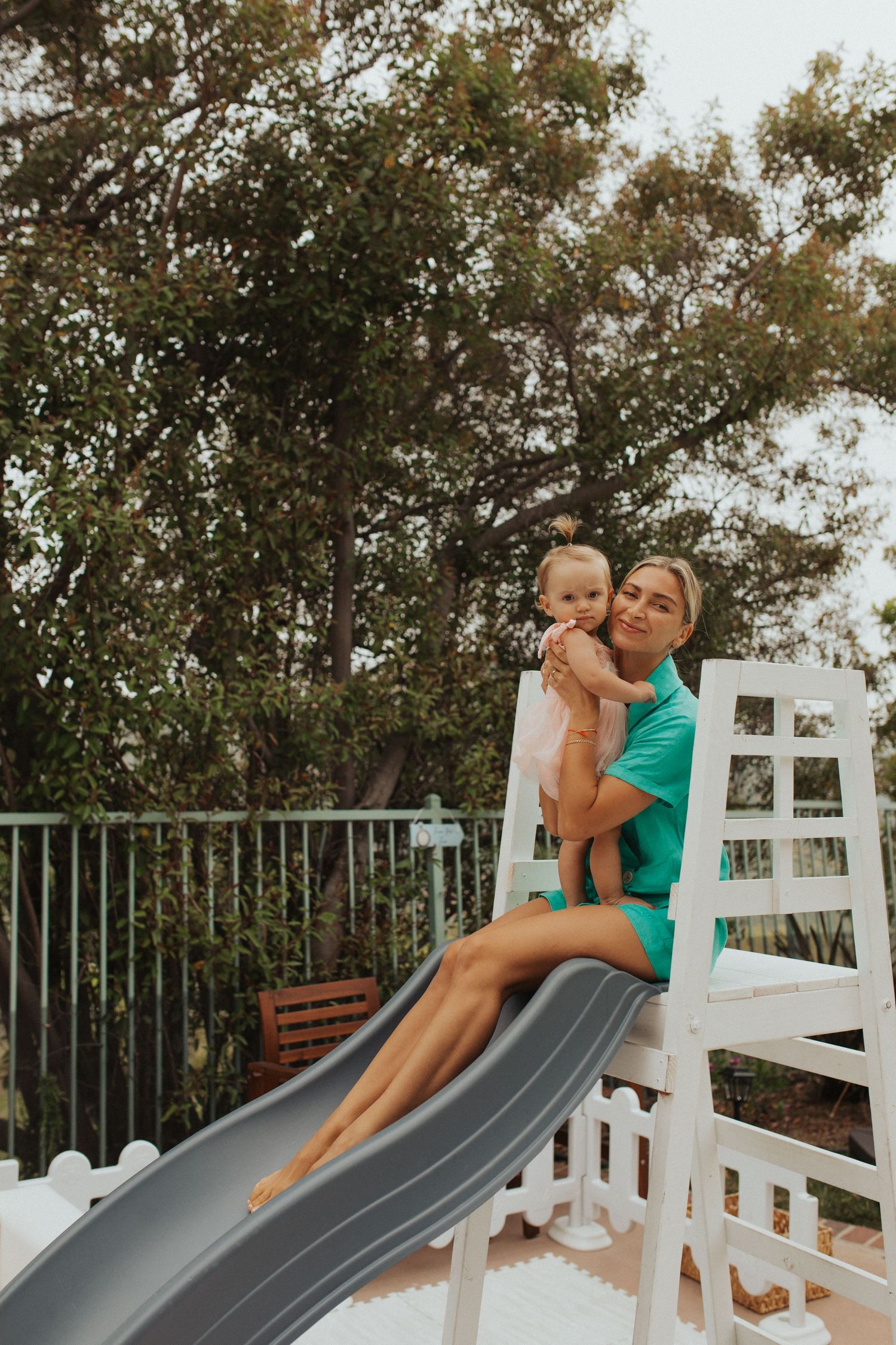 Mother in teal dress holds baby on white playground slide outdoors.