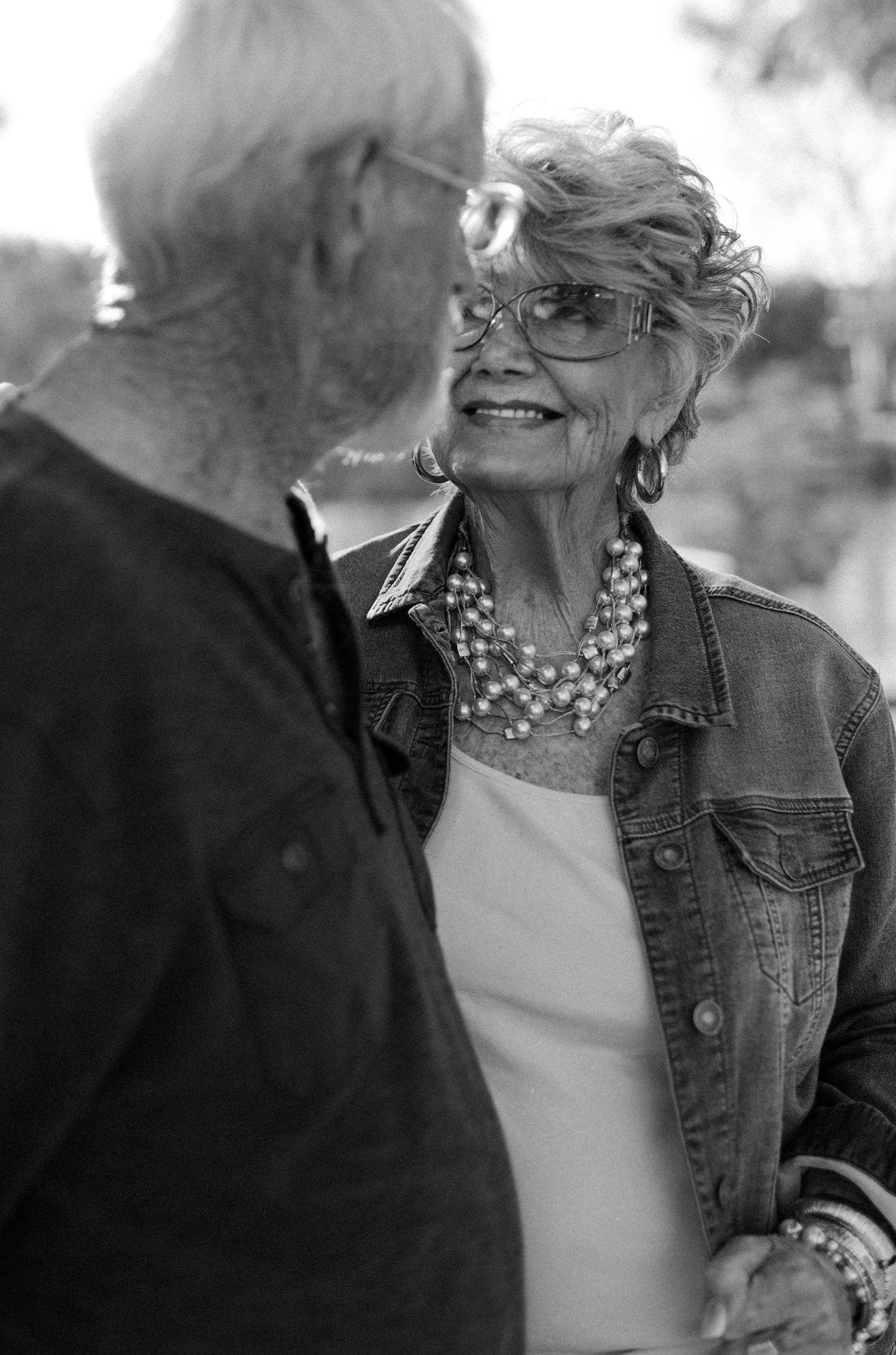 An elegant older woman smiles warmly at her partner in a candid black and white outdoor portrait.