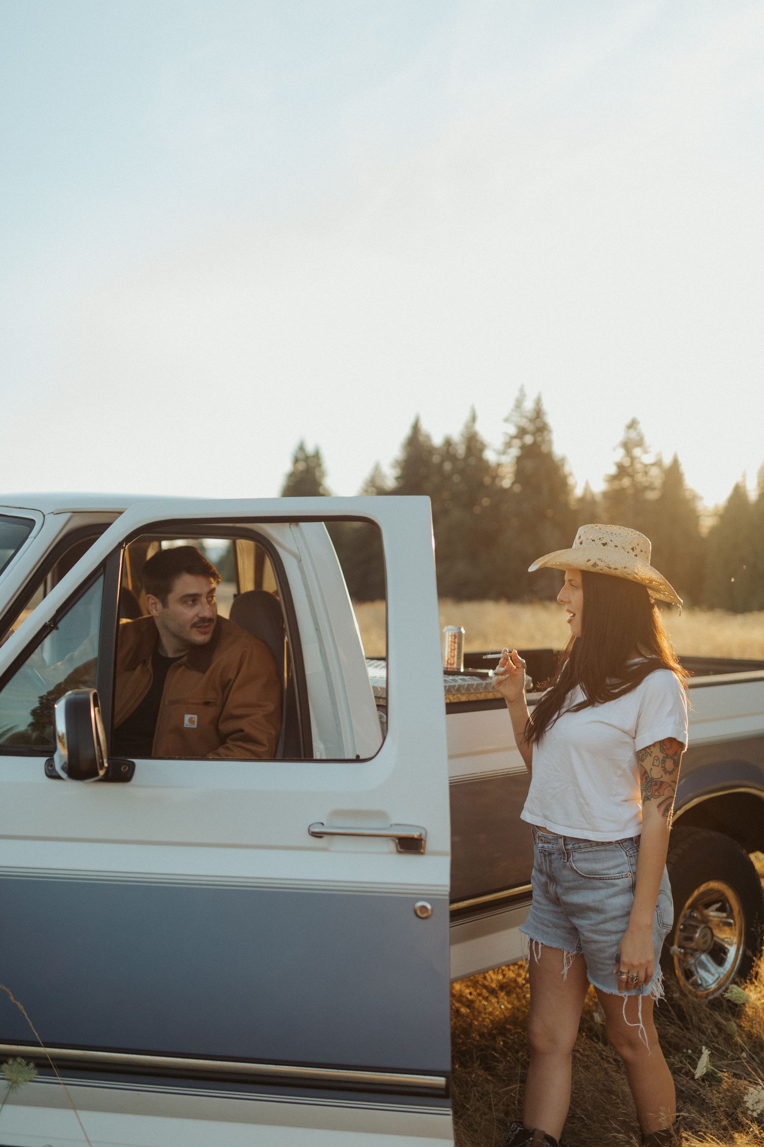 Woman in cowboy hat and denim shorts chatting with man sitting in vintage white pickup truck at sunset.