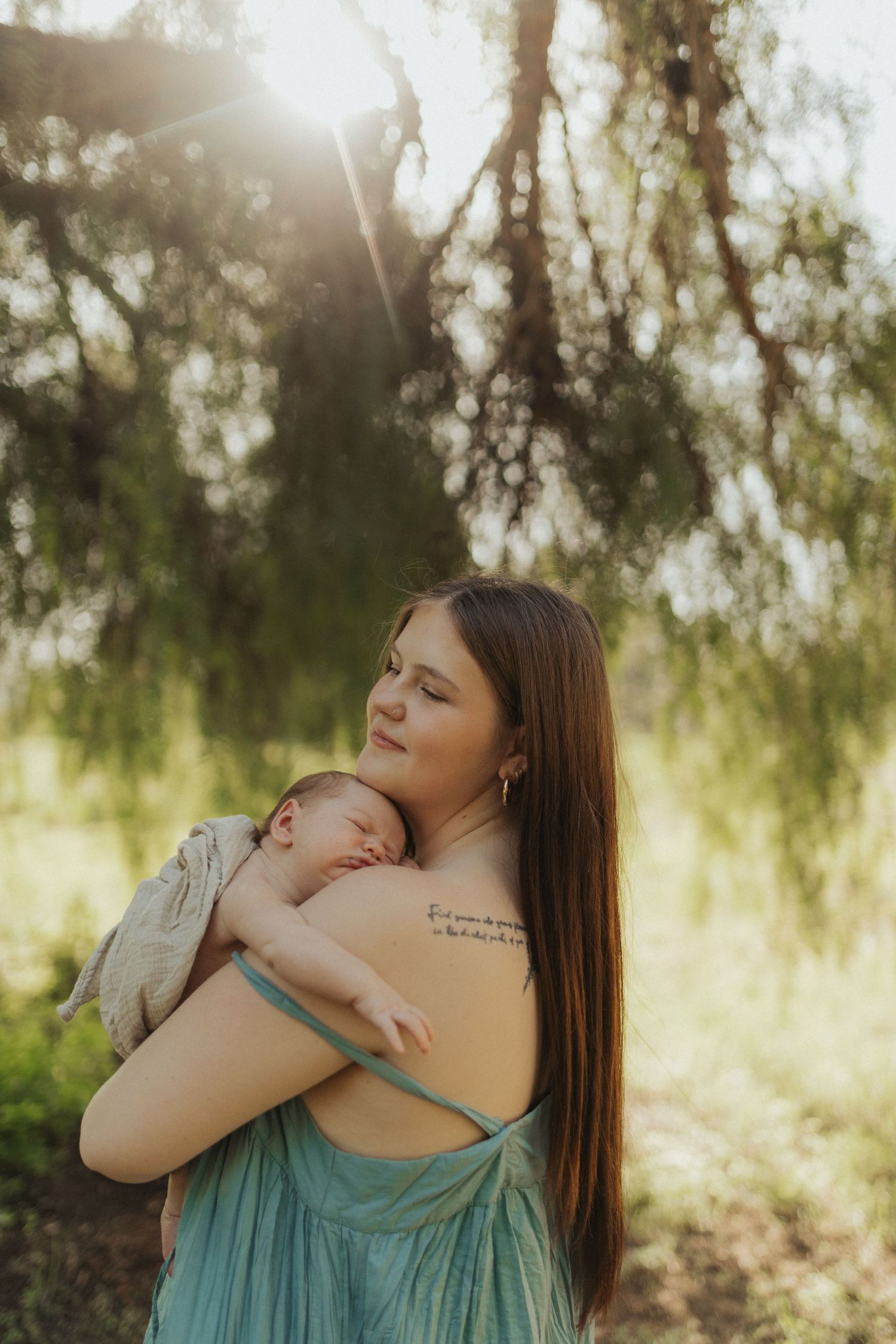 Mother in teal dress holding sleeping newborn in golden sunlight beneath weeping willow tree.