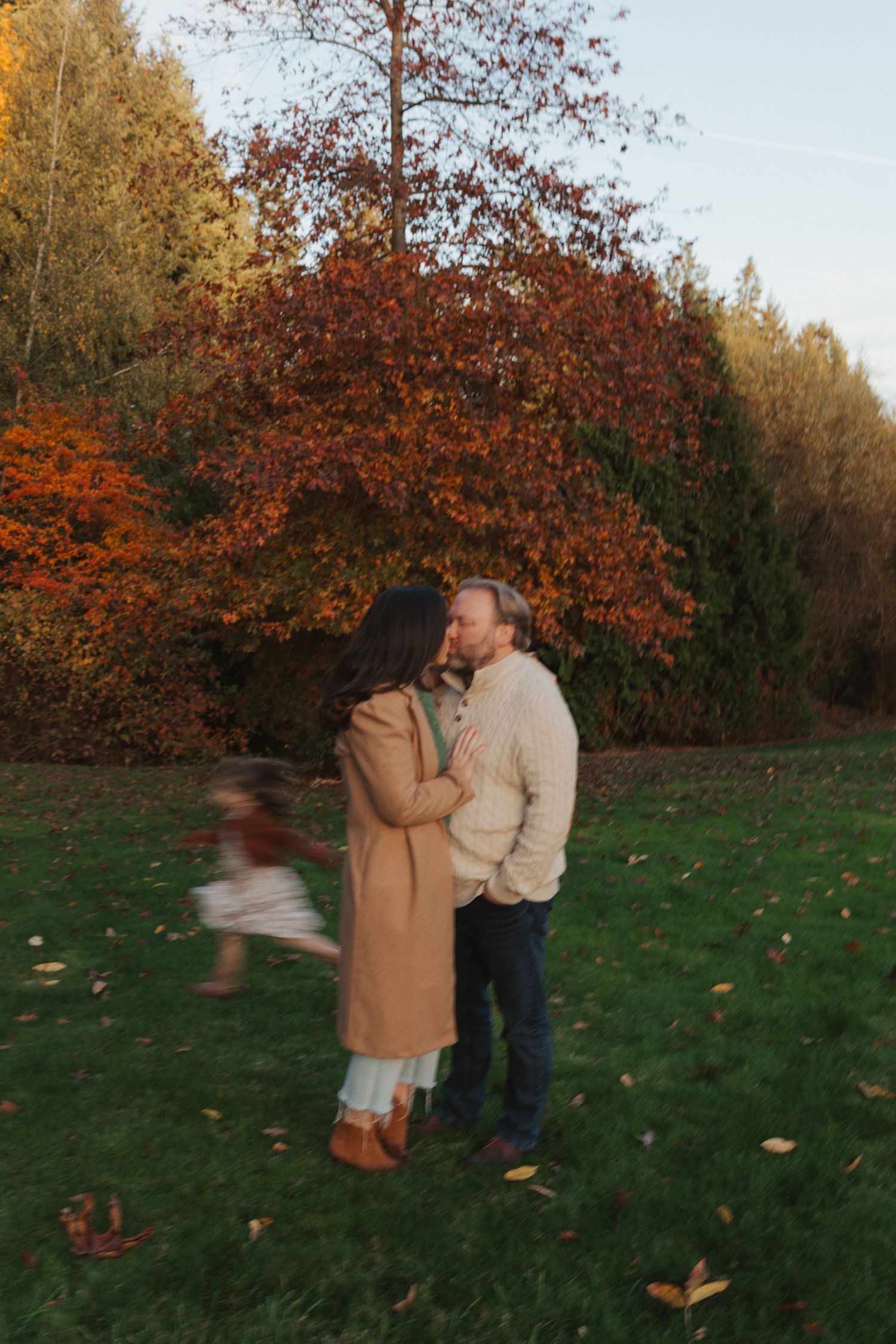 A couple shares a romantic moment on a grass lawn surrounded by stunning red and orange autumn trees.